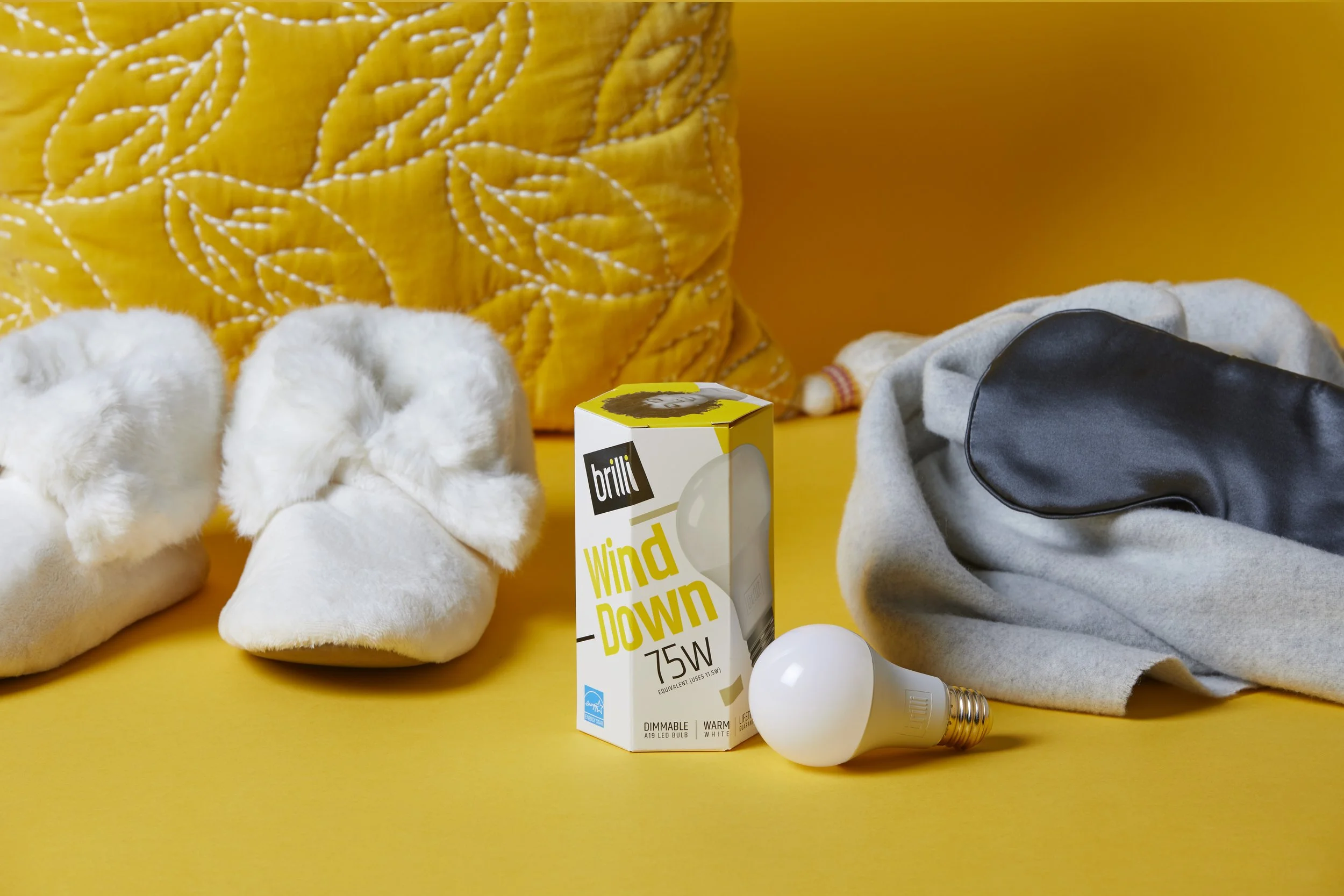 A yellow surface with various objects, including a black and gray purse, a gray cloth, a pair of white fuzzy slippers, a yellow quilted pillow, a lightbulb, and a box of Brill Wind Down 75W light bulb.