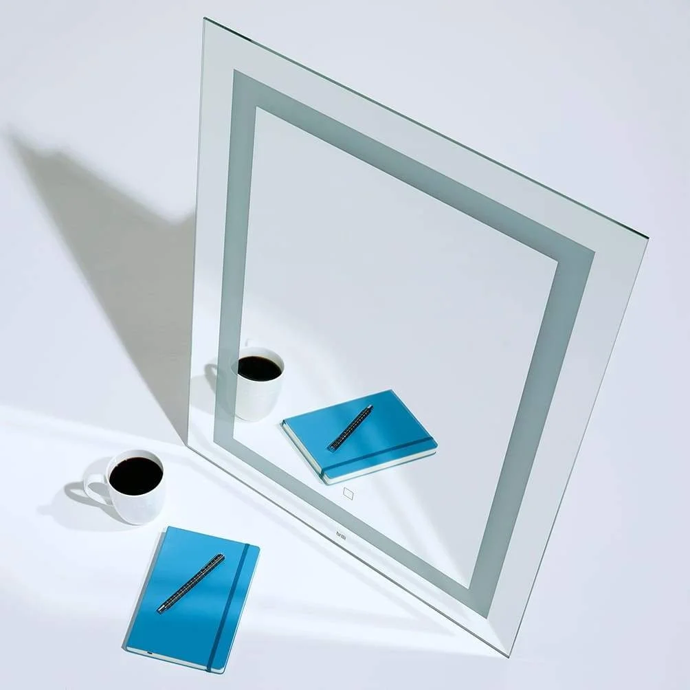 A white square mirror with an open frame standing on a white surface. Two white cups filled with black coffee, two blue notebooks, and one black pen are placed on the surface and reflected in the mirror.