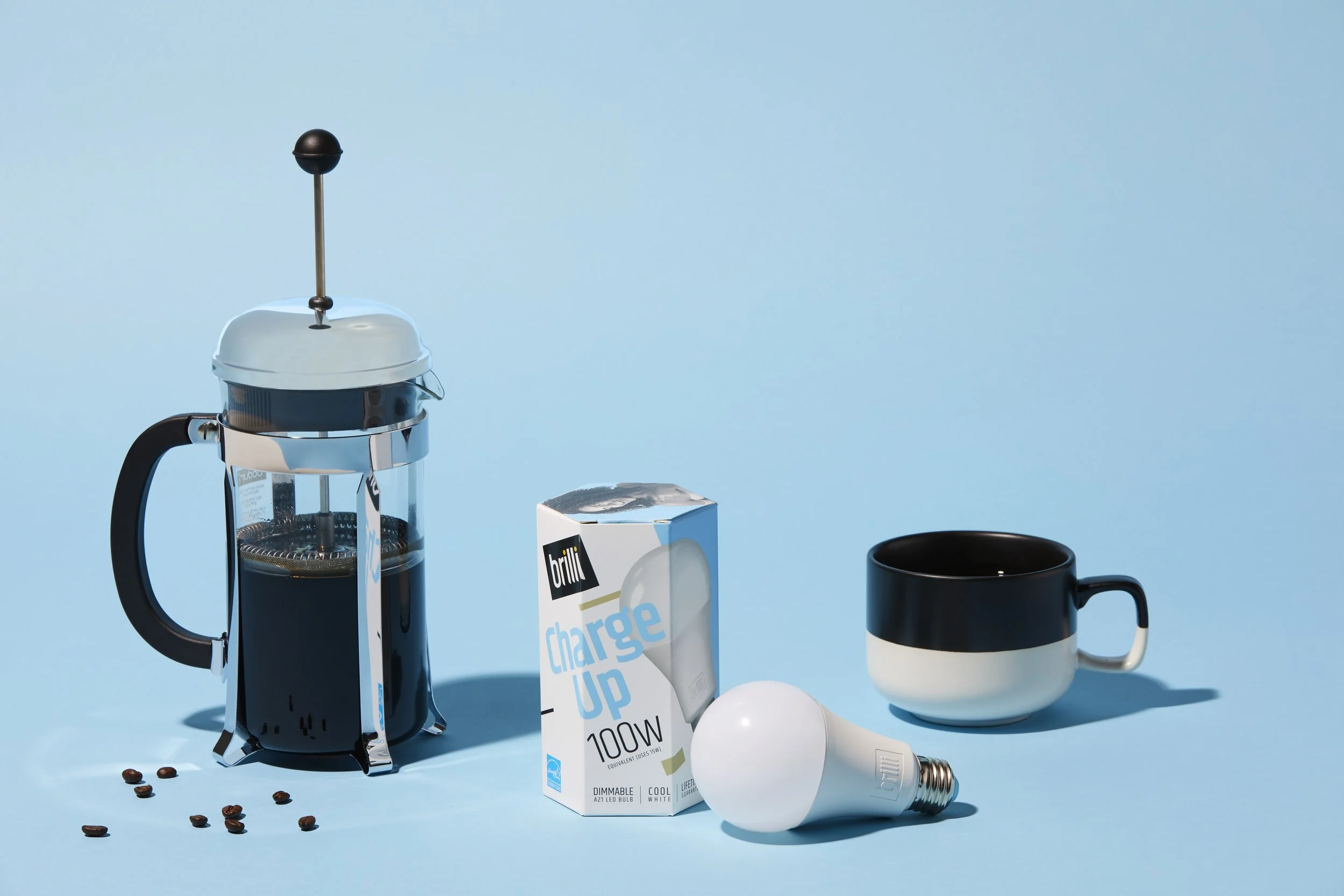 A French press coffee maker with coffee beans, a pack of brilli charge up light bulb, a black and white coffee mug, and a white LED bulb on a light blue background.