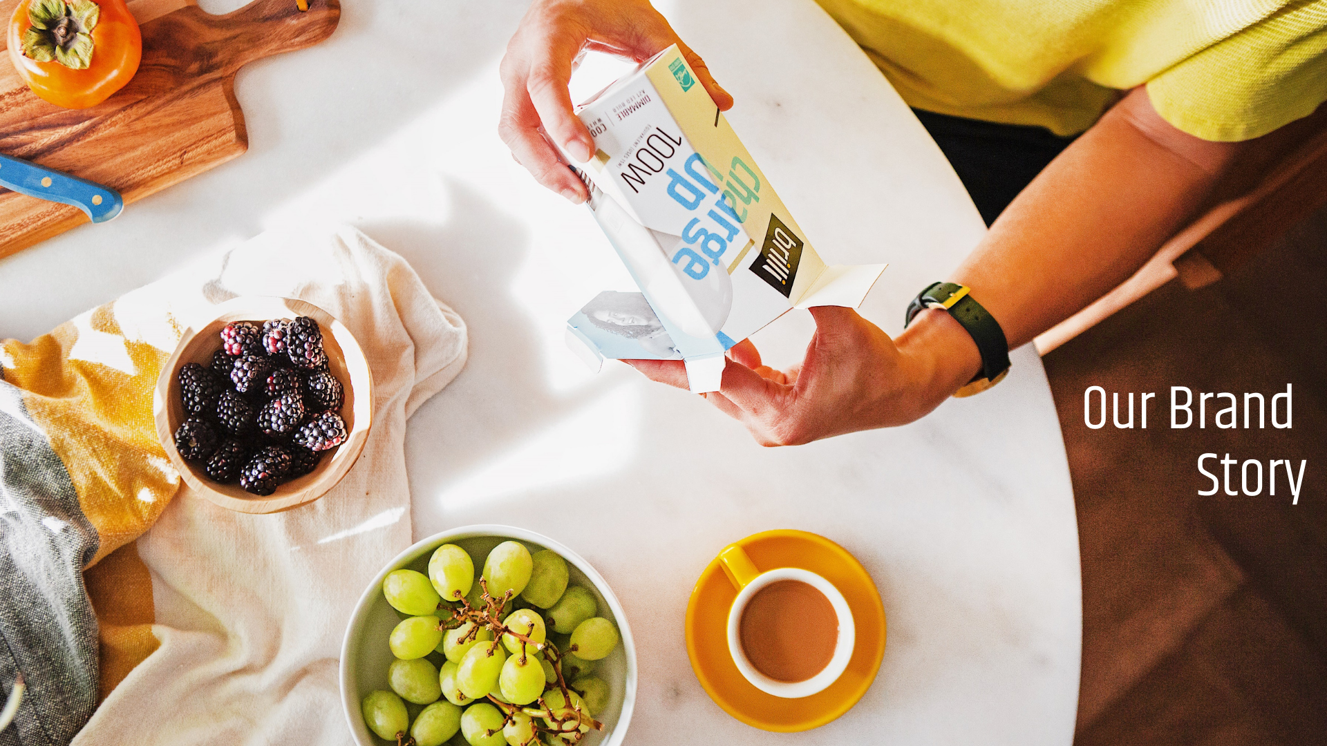 Person pouring almond milk from a carton into a cup on a white table with bowls of blackberries and green grapes, a yellow cloth, and a cutting board with a knife and a persimmon.