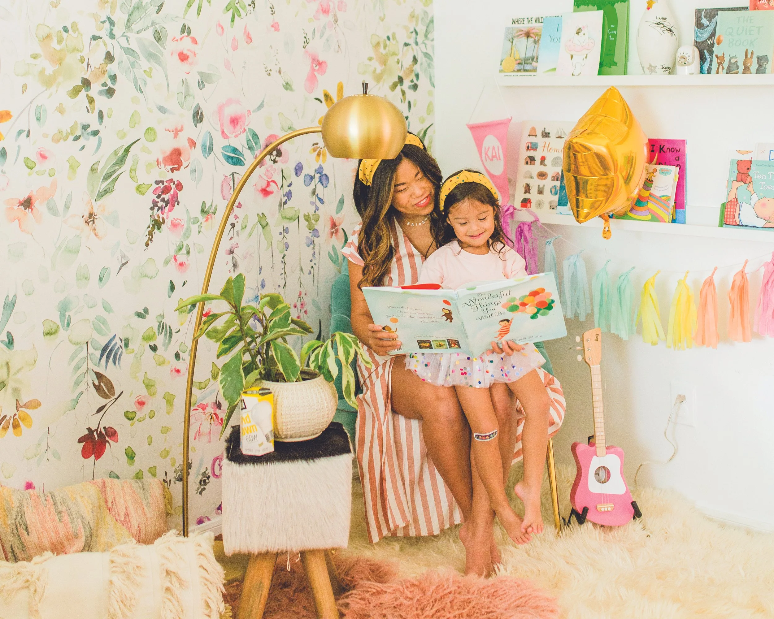 A woman and a young girl sitting together on a chair reading a book in a colorful, decorated room with balloons, books, a guitar, and colorful tassels on the wall.