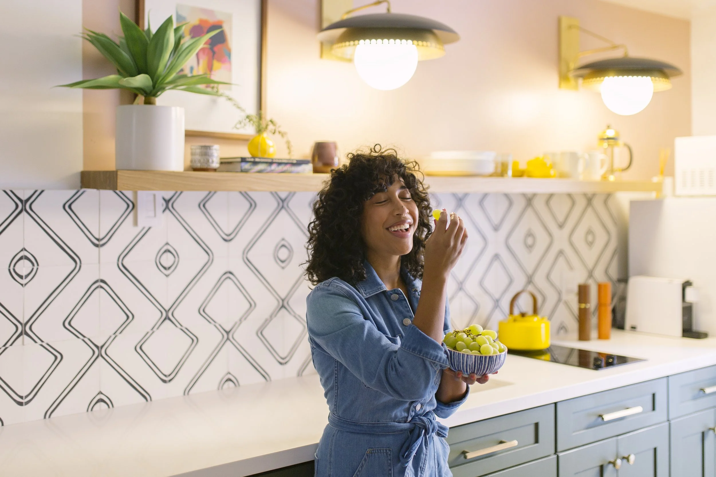 A woman standing in a modern kitchen, holding a bowl of green grapes and smiling while eating one.