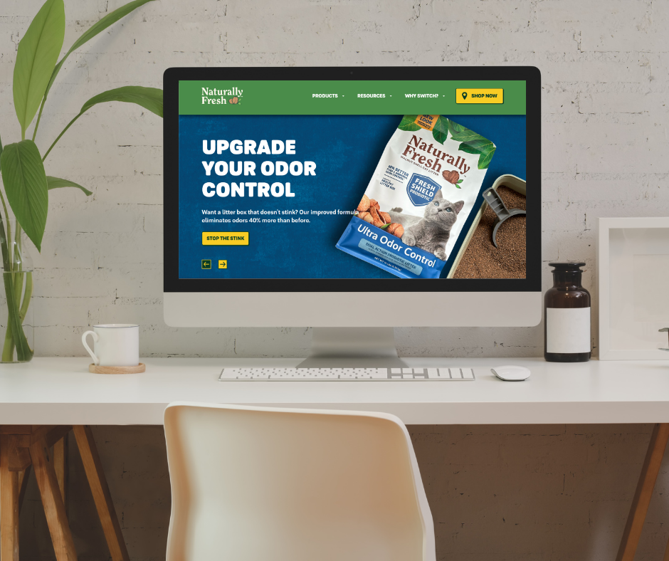 A computer on a white desk displaying a website for Naturally Fresh odor control cat litter with a green header, blue background, and a package of cat litter. The desk has a white mug, a plant, a brown bottle, and a white picture frame. There is a beige chair in front of the desk.
