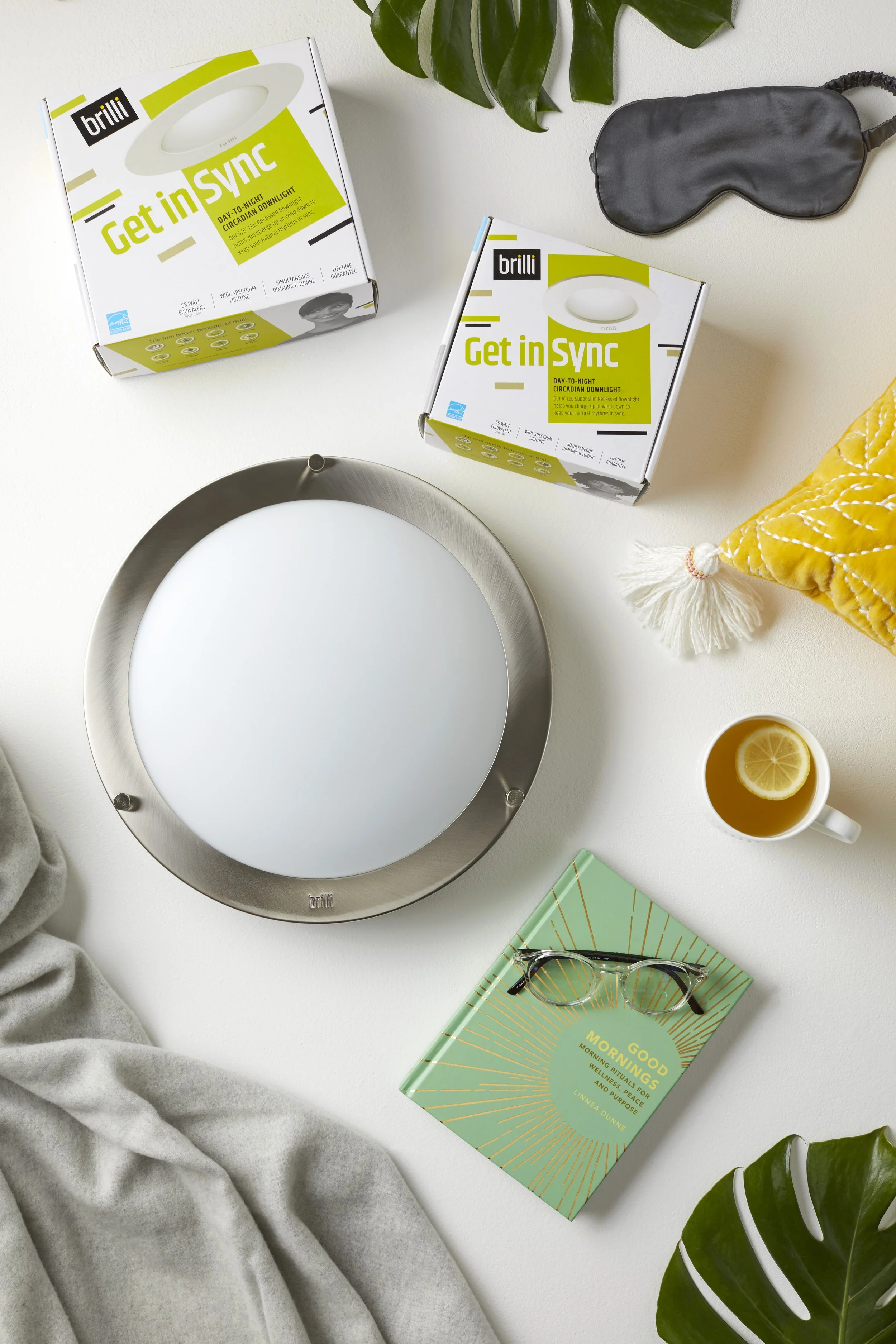A flat lay of modern lifestyle items on a white surface, including a round LED ceiling light, two boxes of 'Get in Sync' circadian nightlights, a black sleep mask, a yellow throw pillow with tassels, a cup of tea with lemon, a book titled 'Good Morni