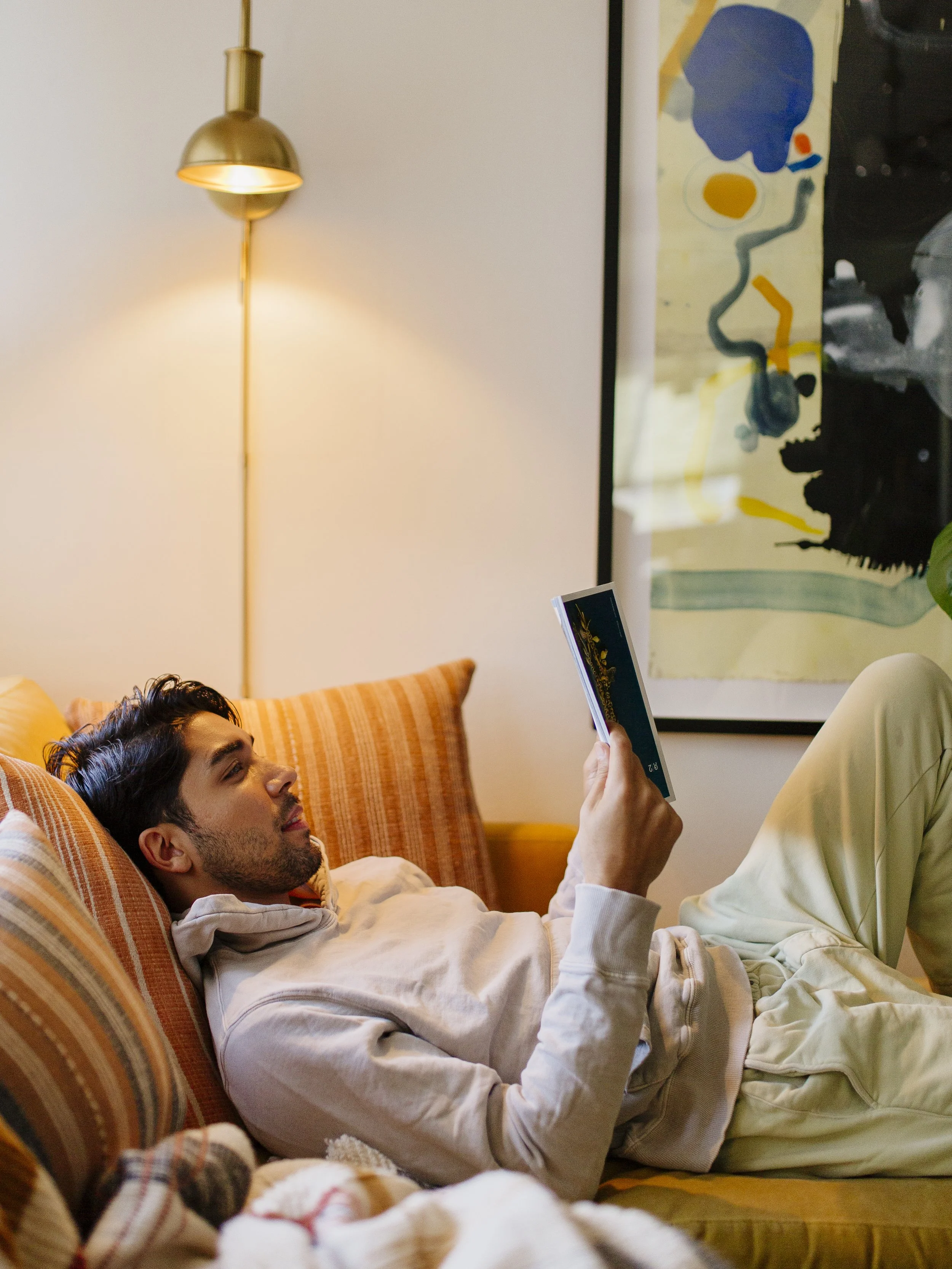 A man is lying on a striped orange couch, reading a book, with colorful cushions and a modern lamp on the wall behind him.