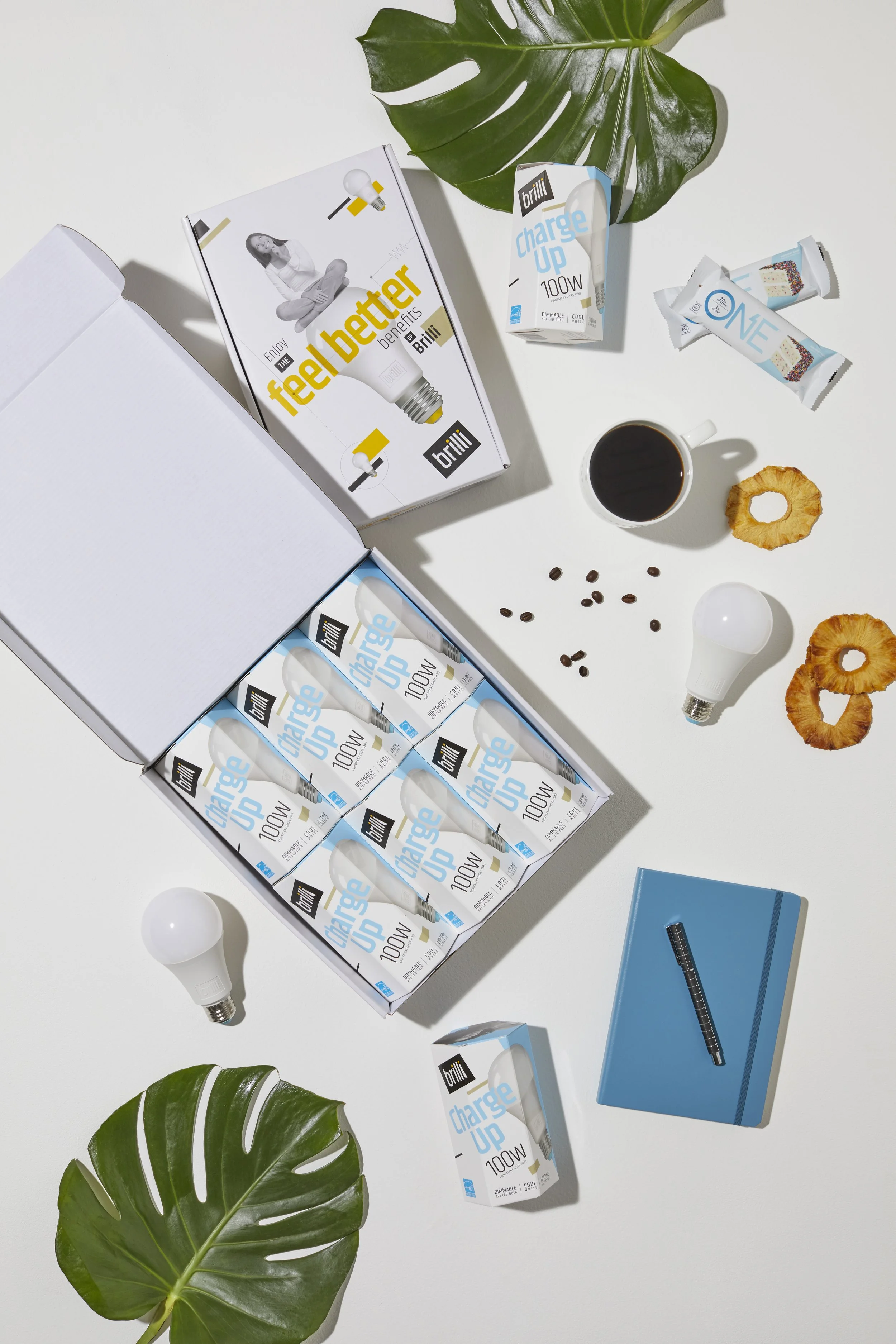 Open white box containing multiple boxes of Brill Charge Up 100W energy-saving light bulbs, placed on a white surface. Various items including coffee, coffee beans, coffee rings, a coffee mug, a small wrapped treat, a headlamp bulb, a large green mon