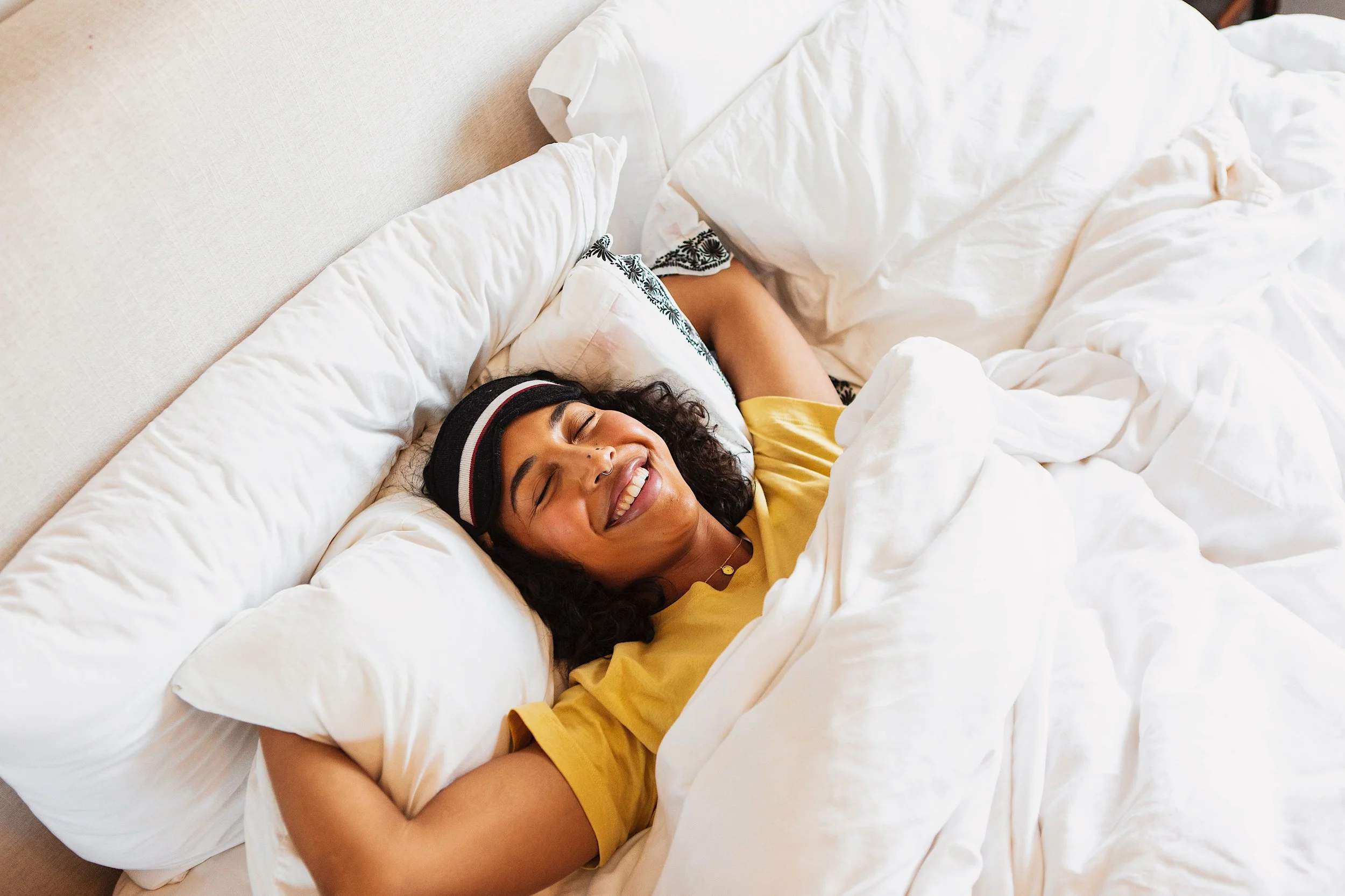 Woman smiling and sleeping on a bed with white pillows and comforter, wearing a yellow shirt and a headband.