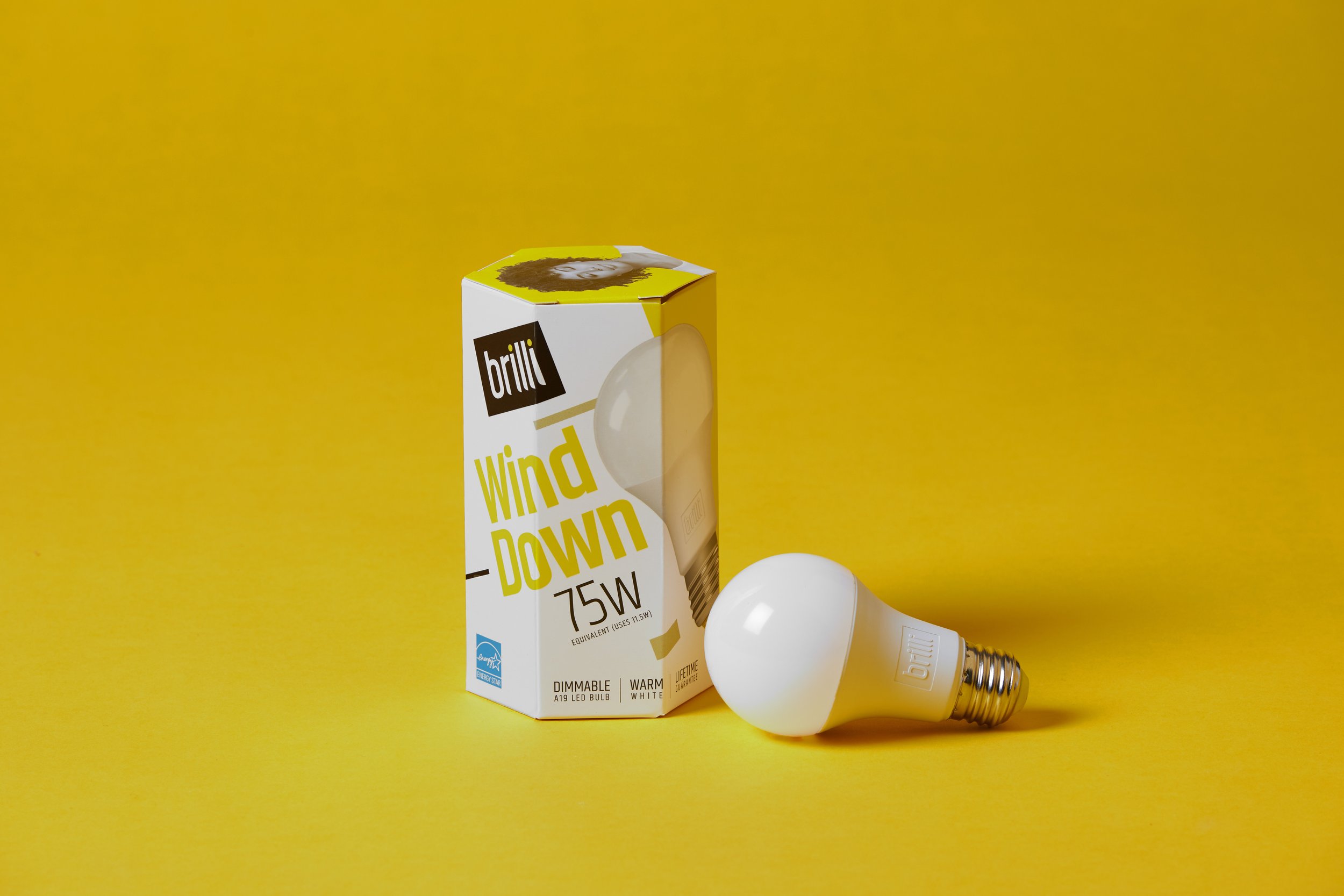 A white LED light bulb placed on a yellow surface next to its packaging box labeled 'brilli Wind Down 75W,' featuring a design with a white background, black and yellow text, and an image of the bulb. The packaging indicates the bulb is dimmable, war