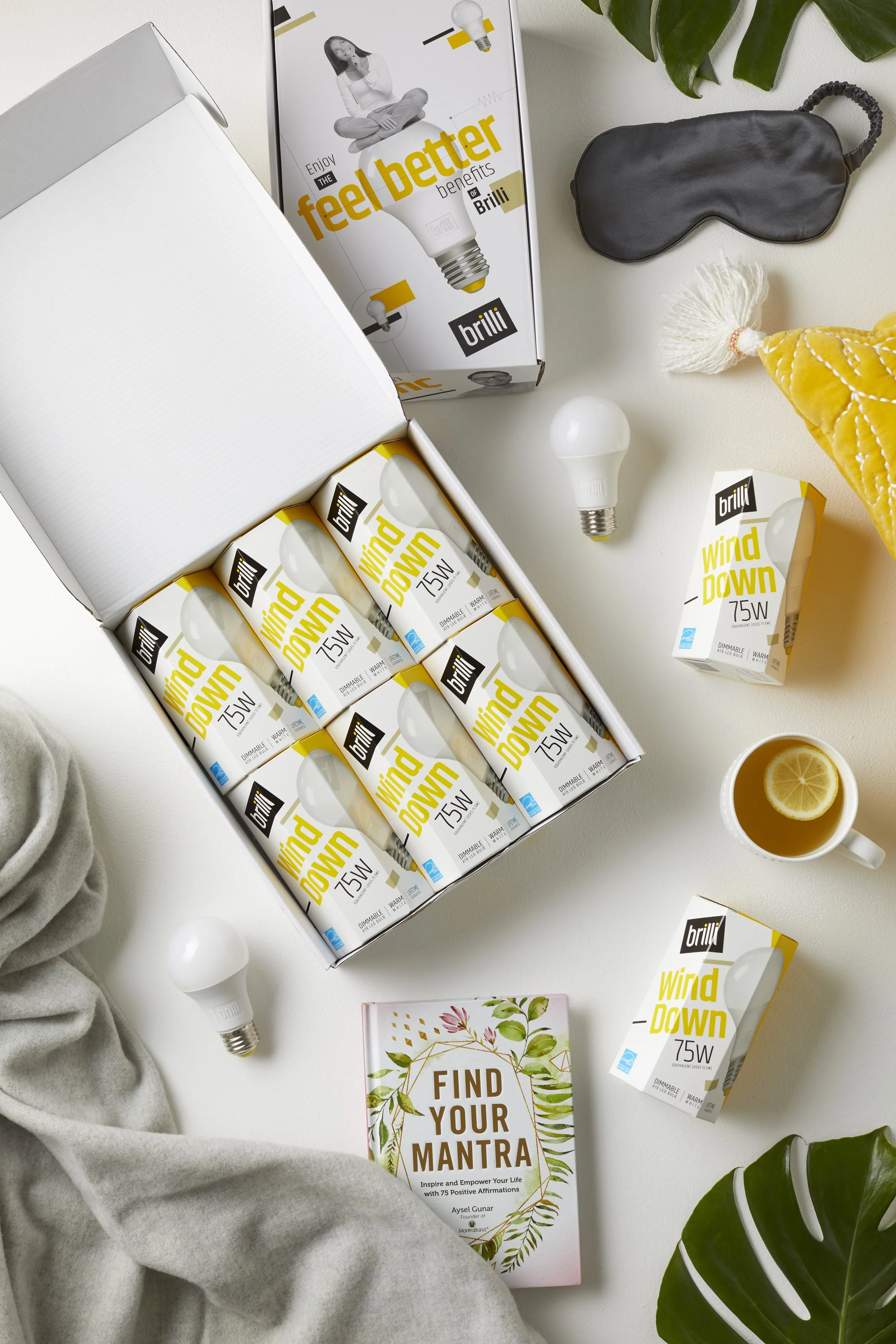 Open box of Brilli Wind Down 75W light bulbs and two additional boxes of the same bulbs on a white table. Nearby are a black sleep mask, a mug with lemon water, a gray blanket, a book titled 'Find Your Mantra', a leafy plant, and a yellow knit hat.