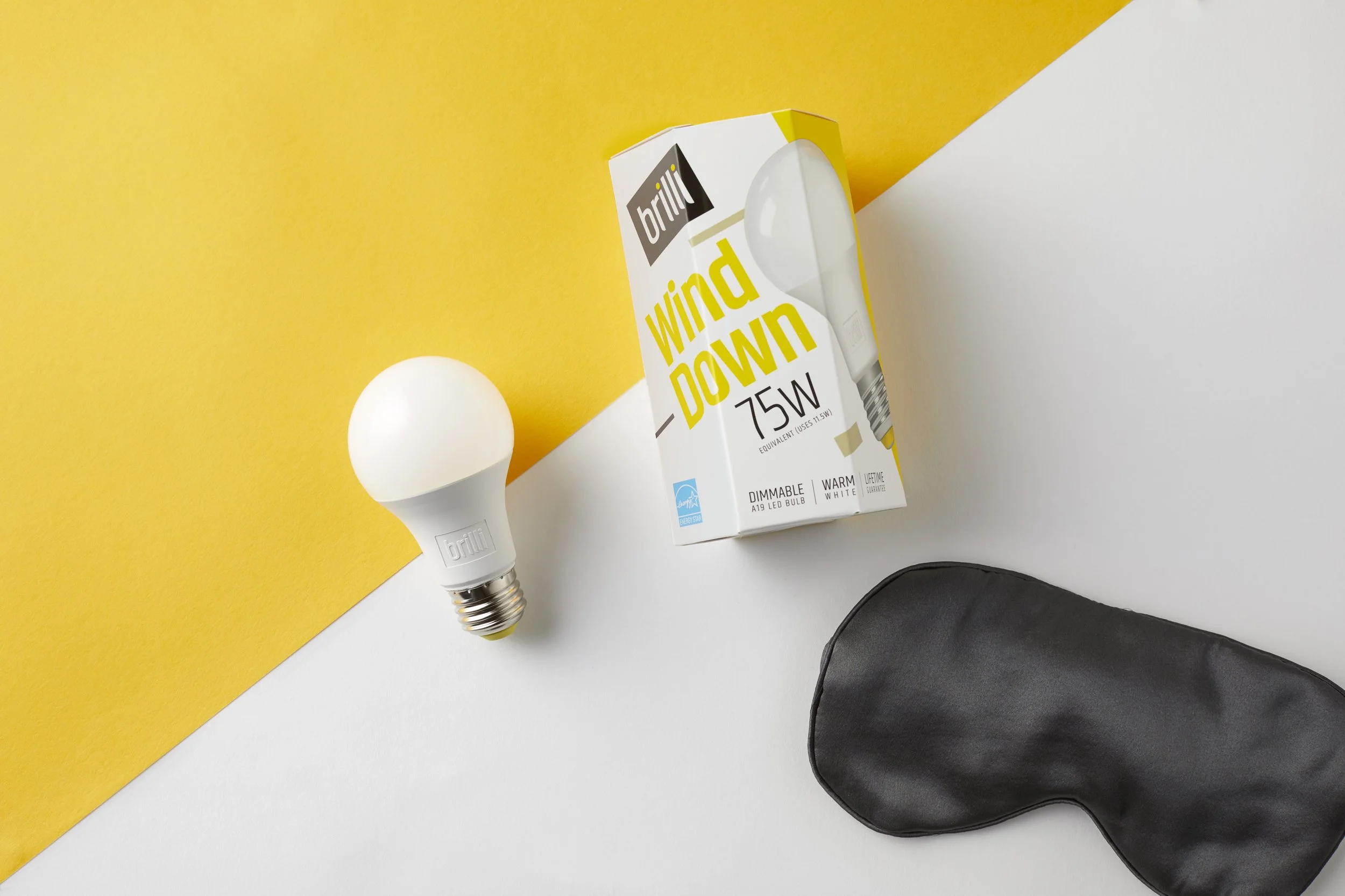 A white smart LED light bulb next to its packaging labeled "Wind Down 75W" on a yellow and white background, and a black sleep mask.