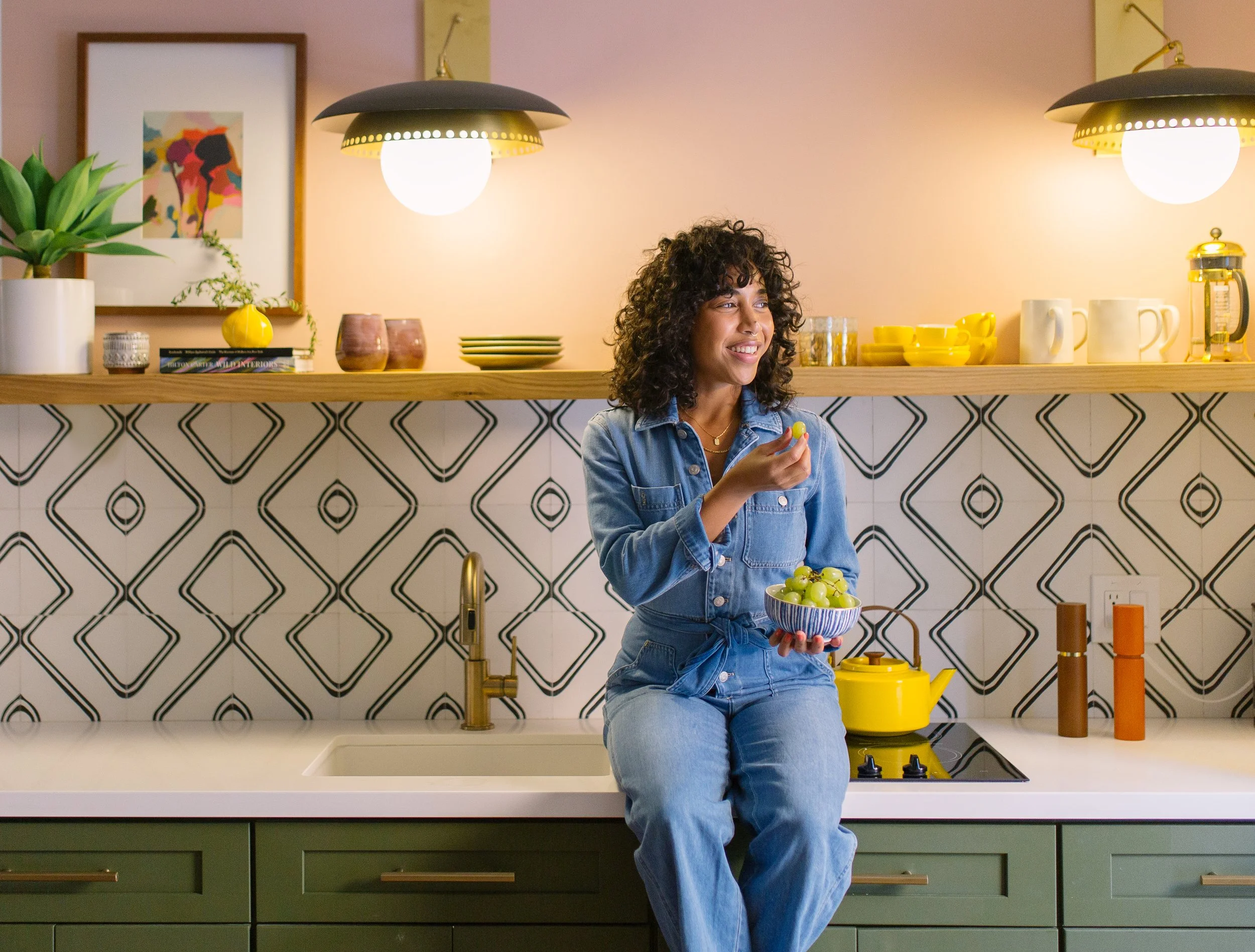 A woman with curly hair in a denim outfit sitting on a kitchen counter holding a bowl of green grapes and smiling, with a kitchen backsplash, plants, and kitchenware in the background.