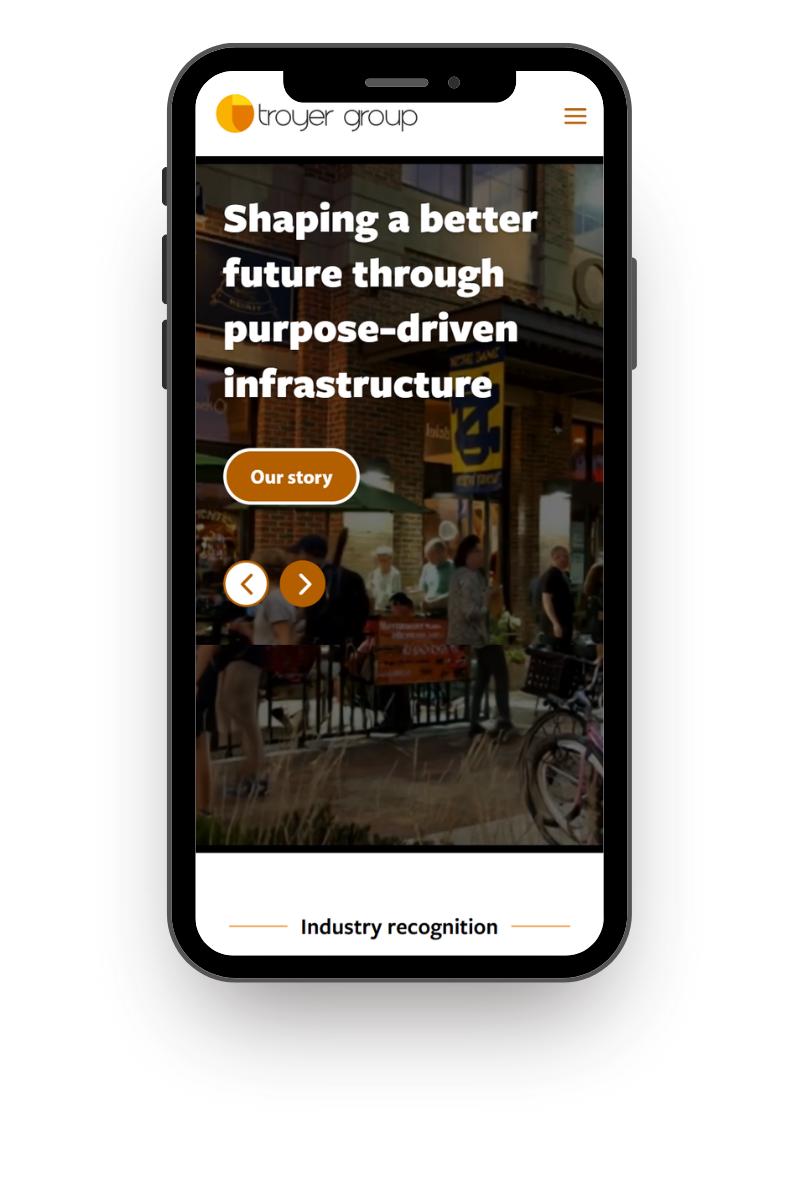 Mobile phone displaying a website for Troyer Group with the headline "Shaping a better future through purpose-driven infrastructure" and a button labeled "Our story" over a background image of an outdoor shopping area with people, brick buildings, and bicycles.