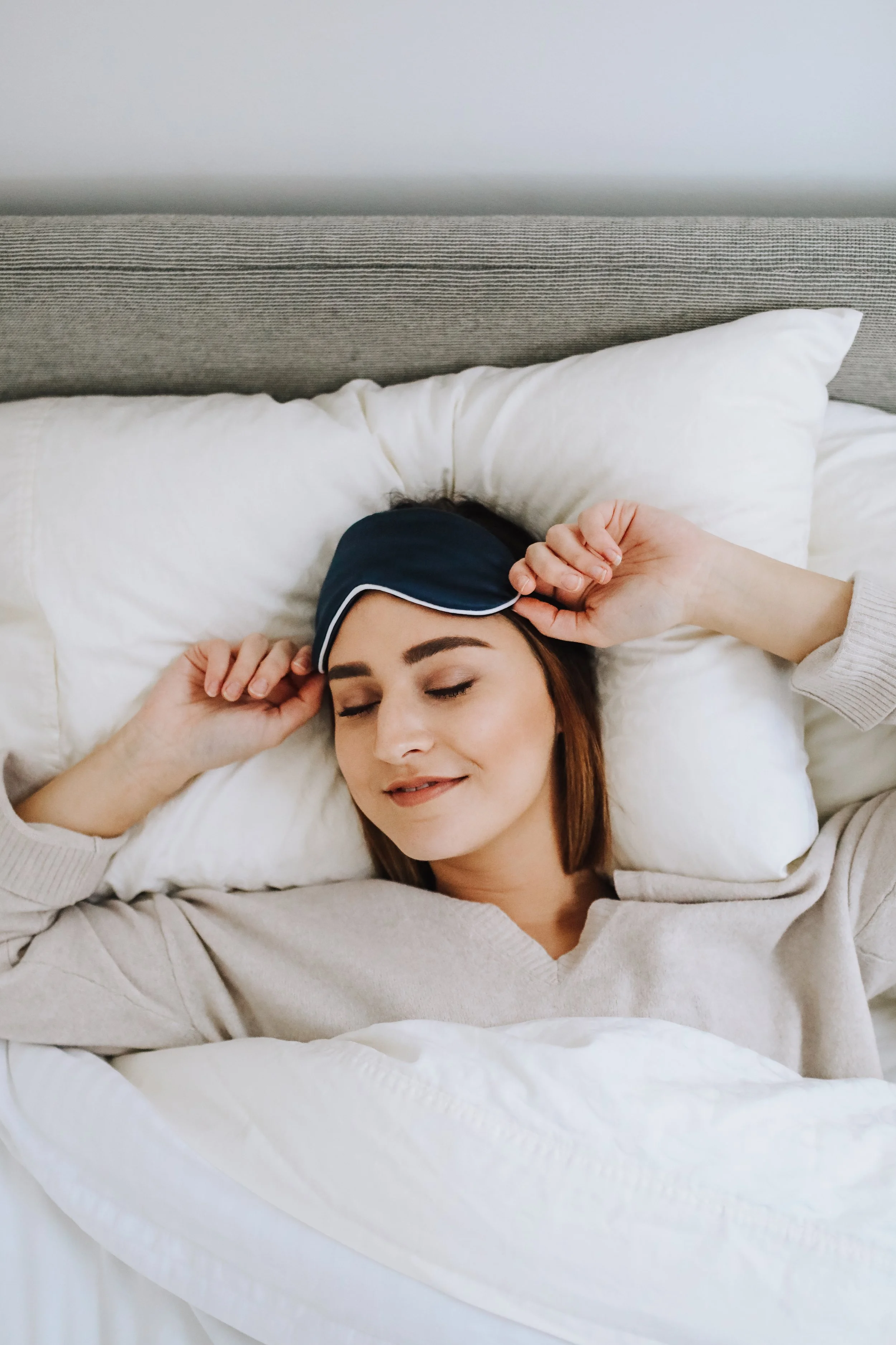 Woman sleeping on her back with a sleep mask on her forehead, smiling with eyes closed, in bed with white bedding and pillows.