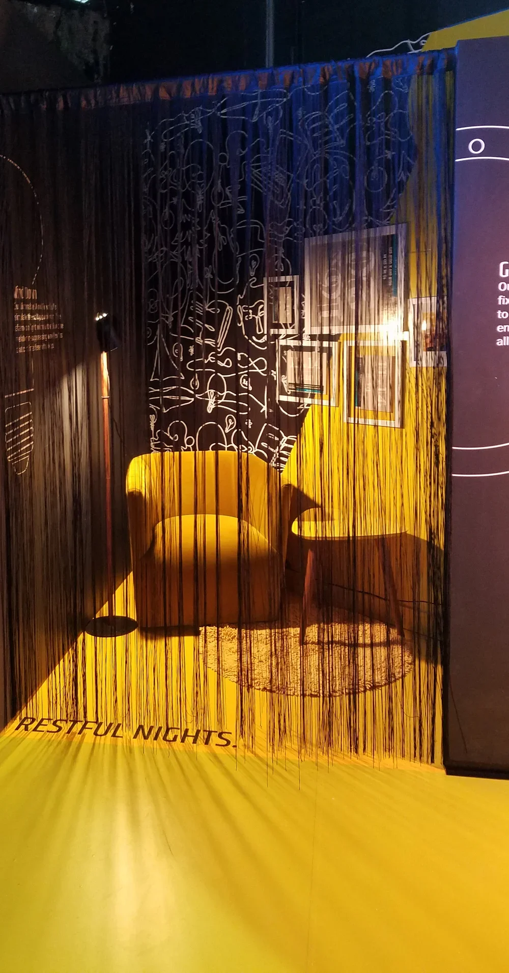 A cozy corner with yellow chairs, a small table, decorative wall art, and a yellow floor mat with the words "Restful Nights" written on it, viewed through black curtain strings.