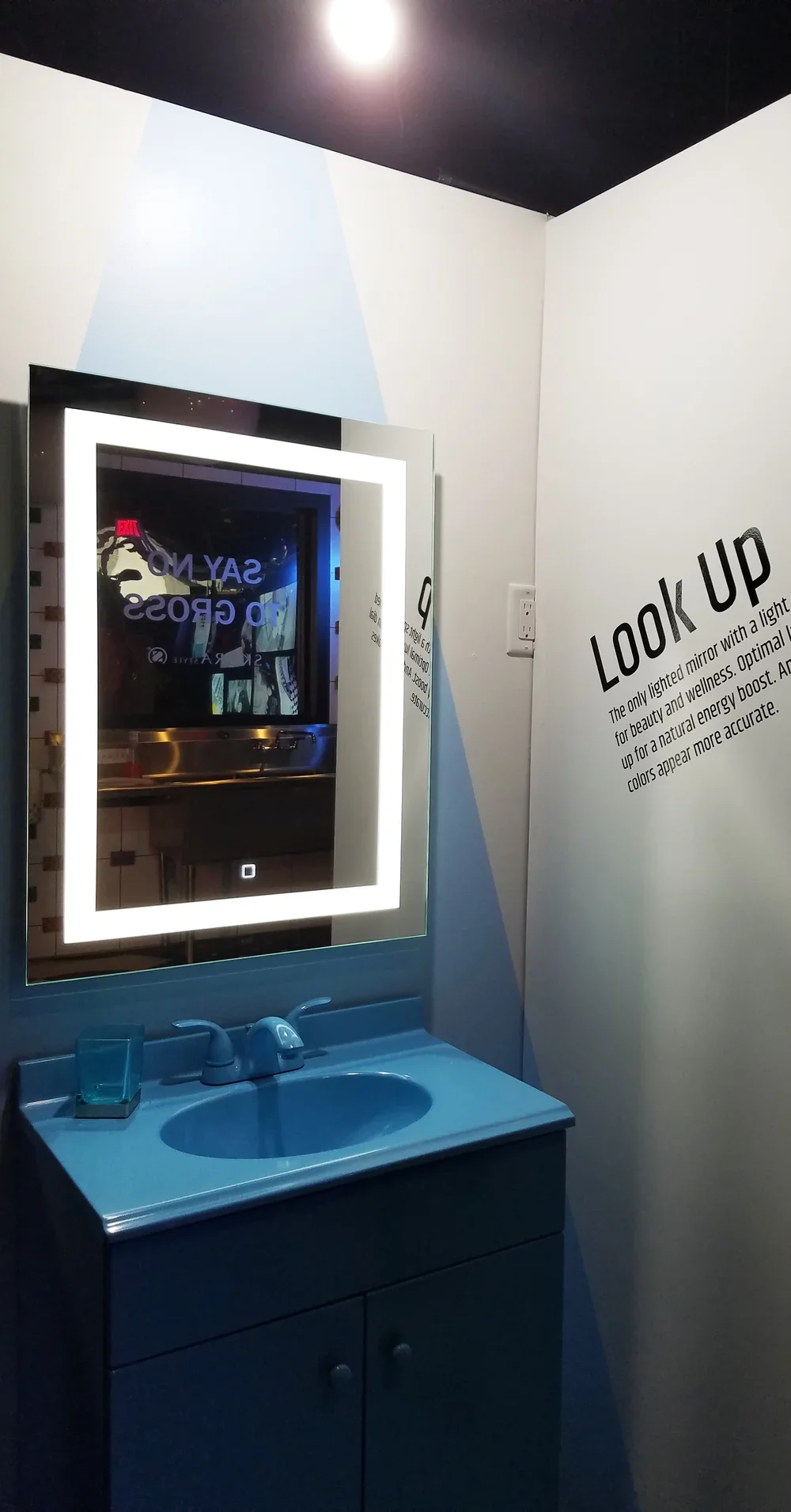 A blue bathroom vanity with a sink, a mirror with LED lighting, and a sign on the wall with instructions to look up for better lighting conditions.