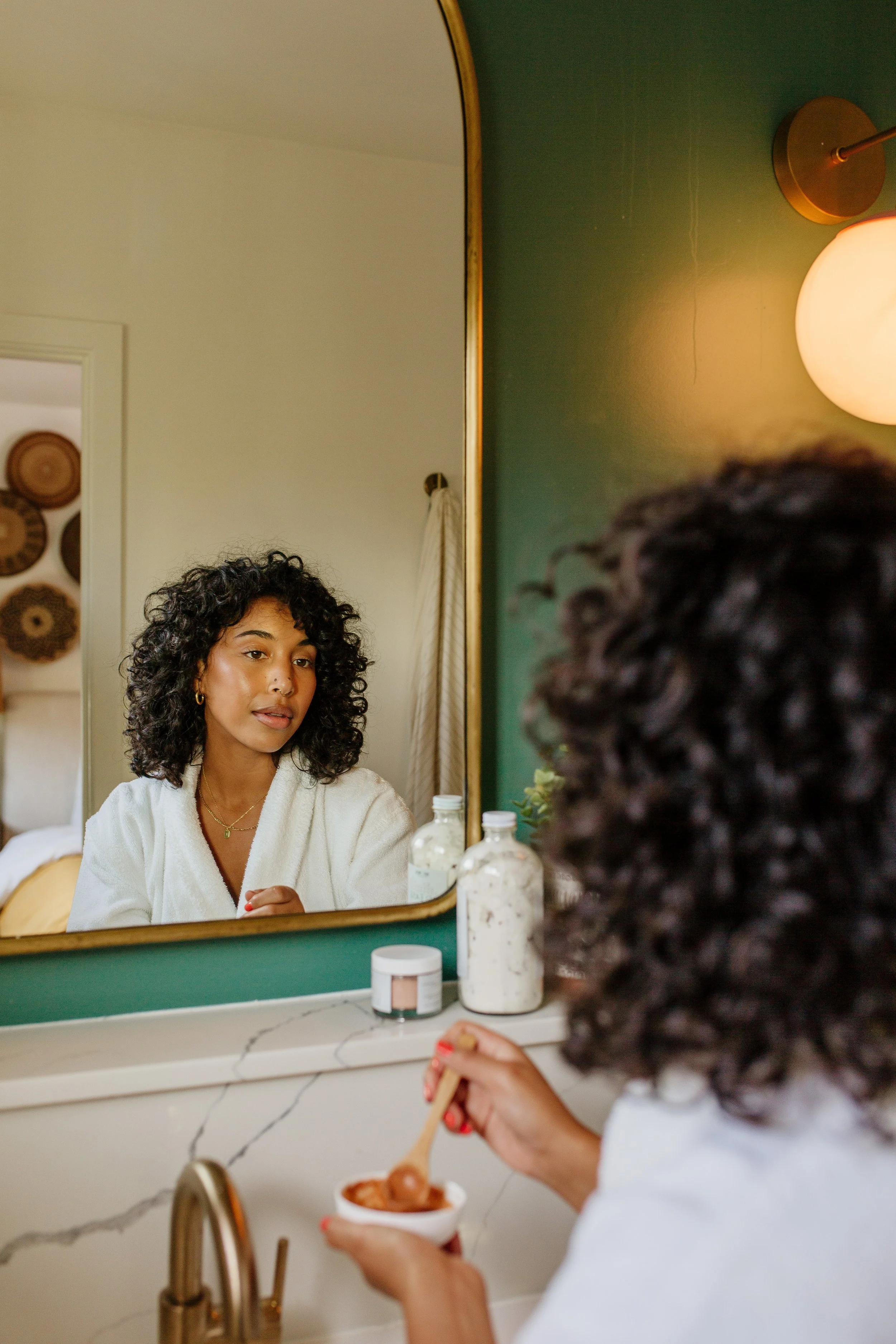 A woman with curly dark hair looking at her reflection in a bathroom mirror while eating food from a bowl with a spoon.