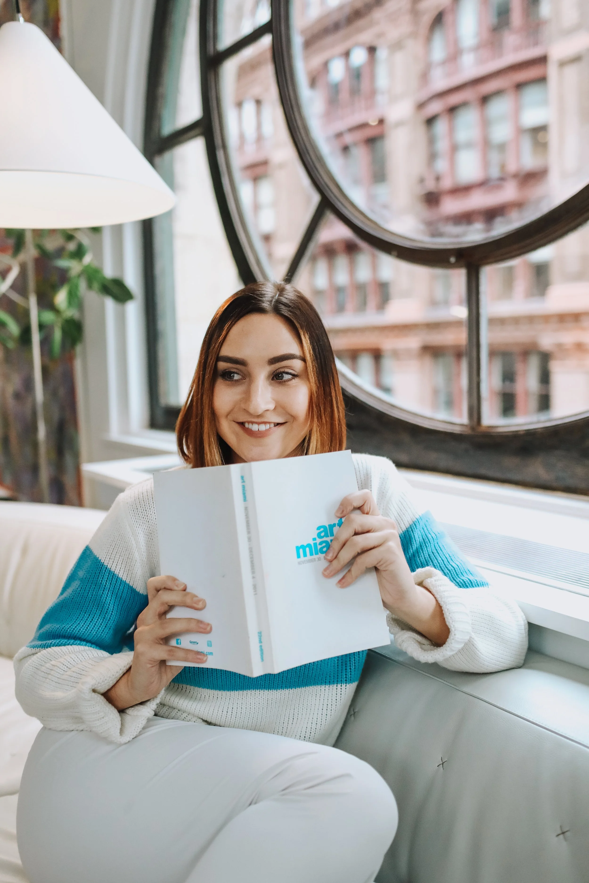 A young woman with shoulder-length brown hair, wearing a white and blue striped sweater, is sitting on a white couch by a window, smiling and holding a booklet titled 'Art Miami' inside a brightly lit room with large round windows and brown brick buildings outside.