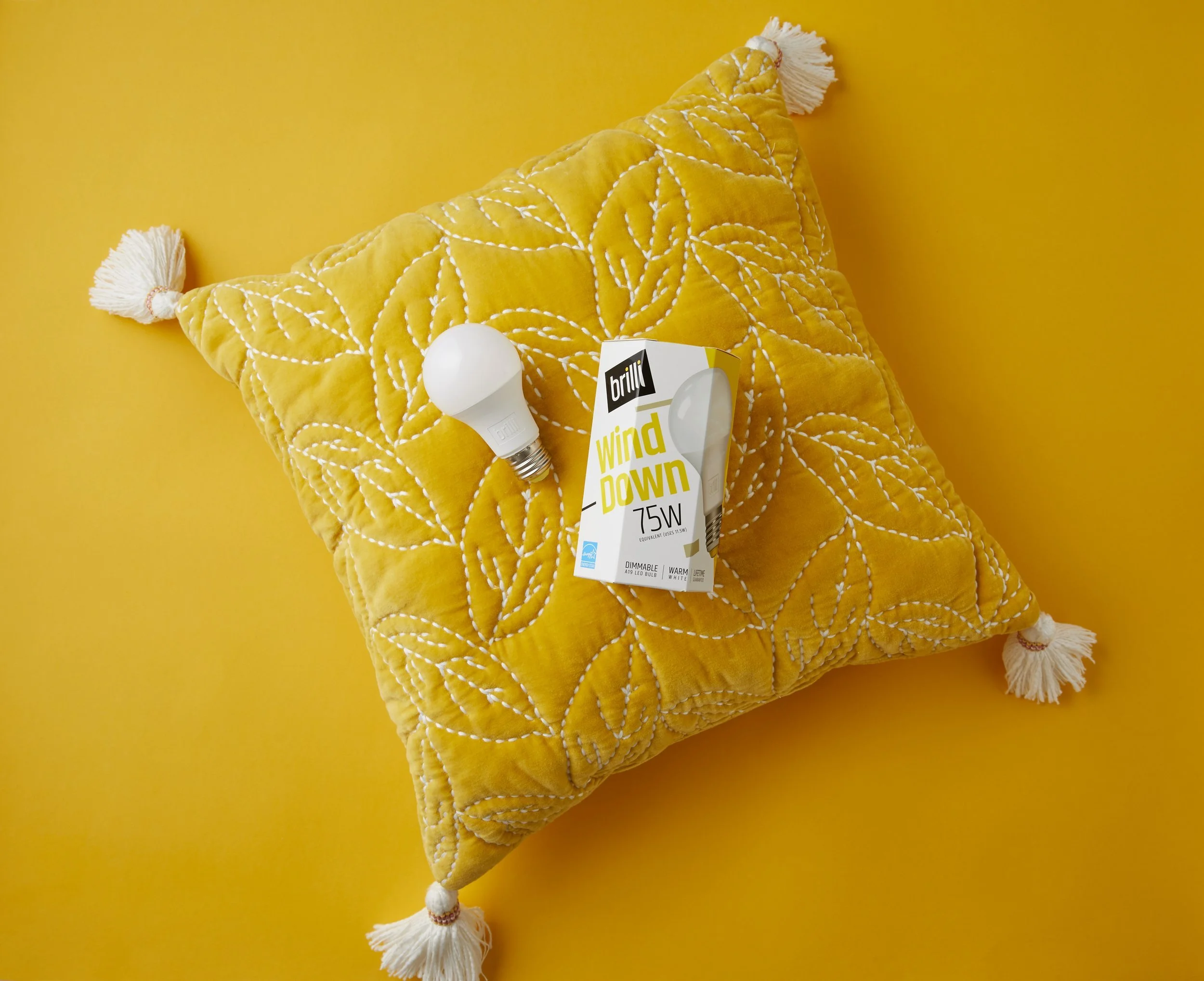 A yellow quilted pillow with white tassels at each corner, placed on a yellow background. On top of the pillow, there is a compact fluorescent light bulb and a box of Brillo wind-down 75W light bulbs.