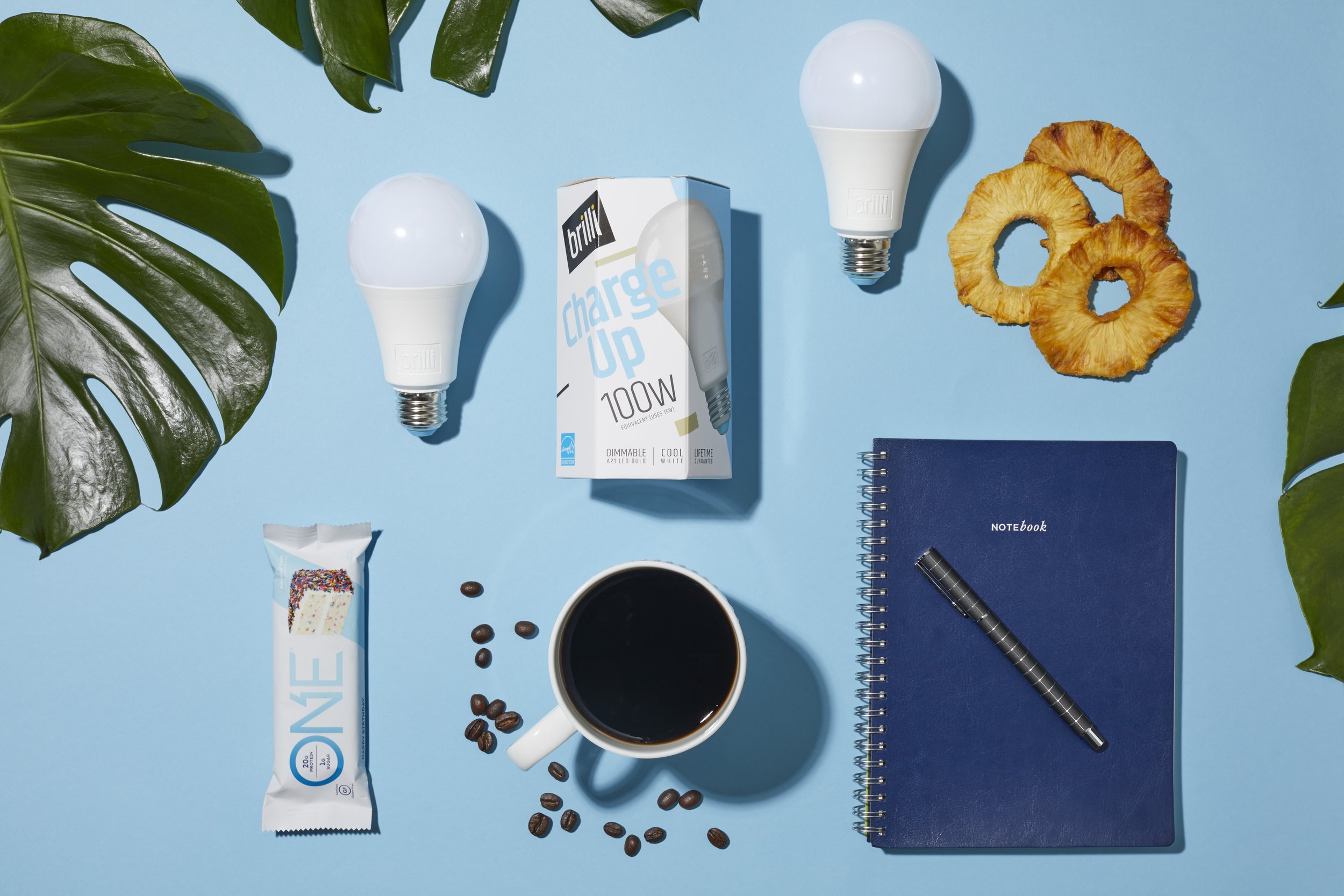 Flat lay of a blue notebook with a black pen on a blue background, surrounded by a cup of coffee, coffee beans, two energy-saving light bulbs, a box of energy-efficient light bulbs, a snack bar, dried pineapple rings, green tropical leaves, and a spiral notebook.