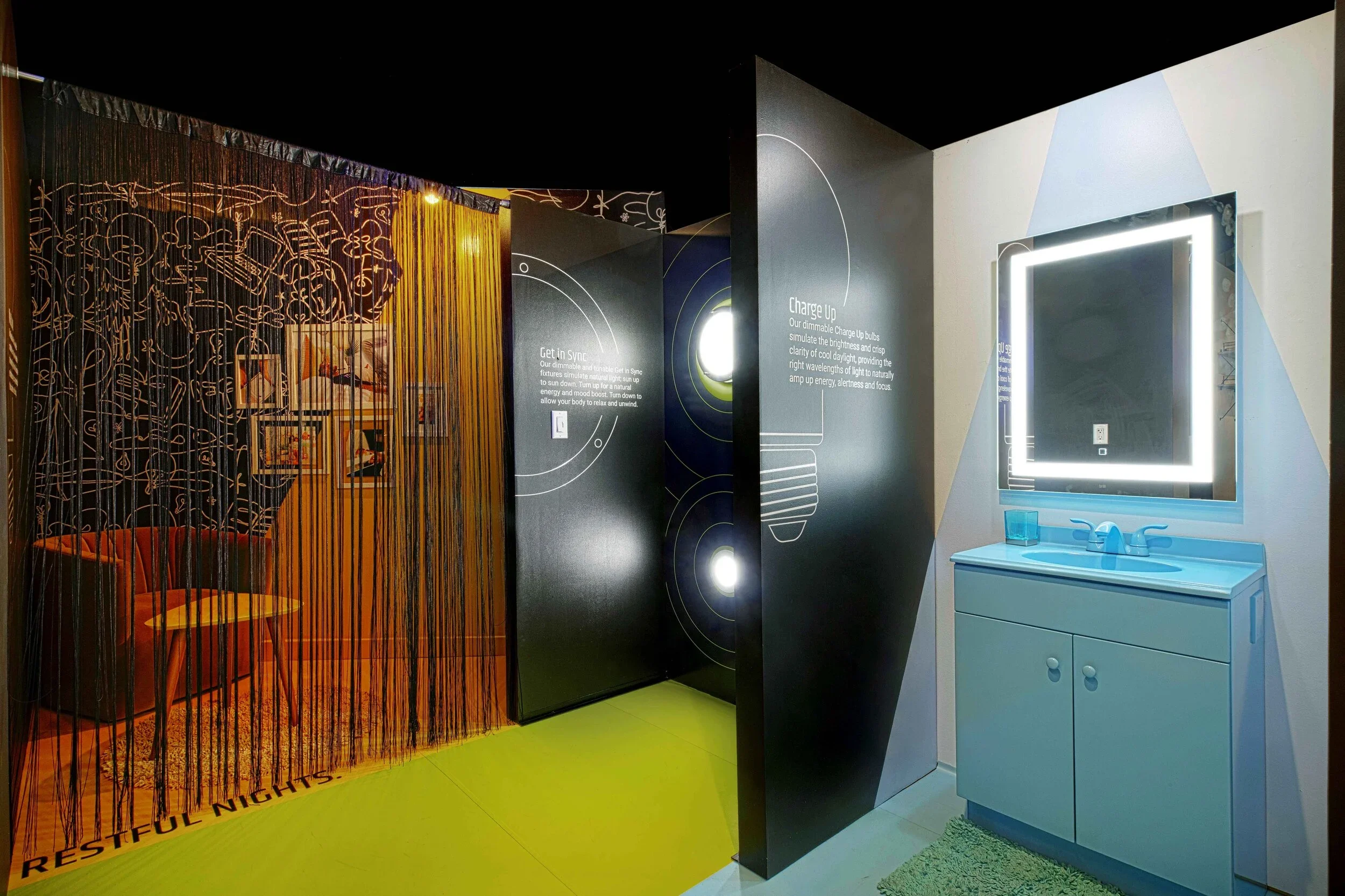 An interactive exhibit with a bathroom scene, including a blue sink and mirror with backlit LED lights, and a black wall with white text explaining charging up lights, connected to a dark room with orange chairs and a wall of artwork behind a string 