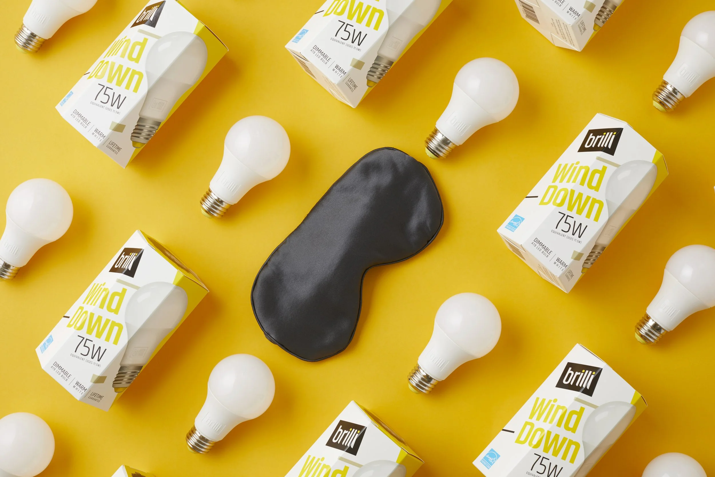 Multiple light bulbs, some in packaging labeled 'brilli Wind Down 75W' and others unpackaged, surrounding a black sleep mask on a yellow background.