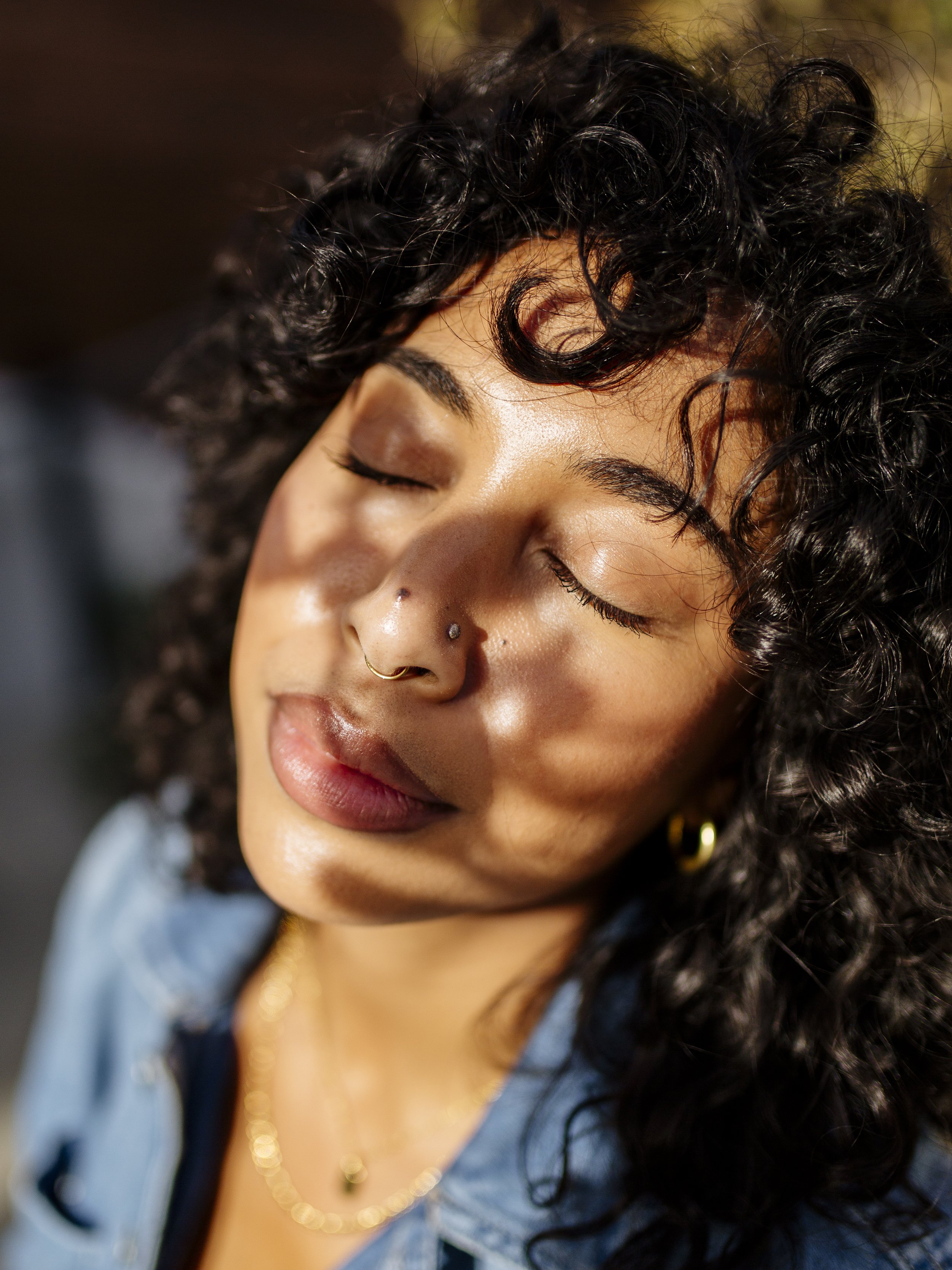 Close-up of a young woman with curly hair, closed eyes, and a relaxed expression, wearing gold jewelry, in warm lighting.