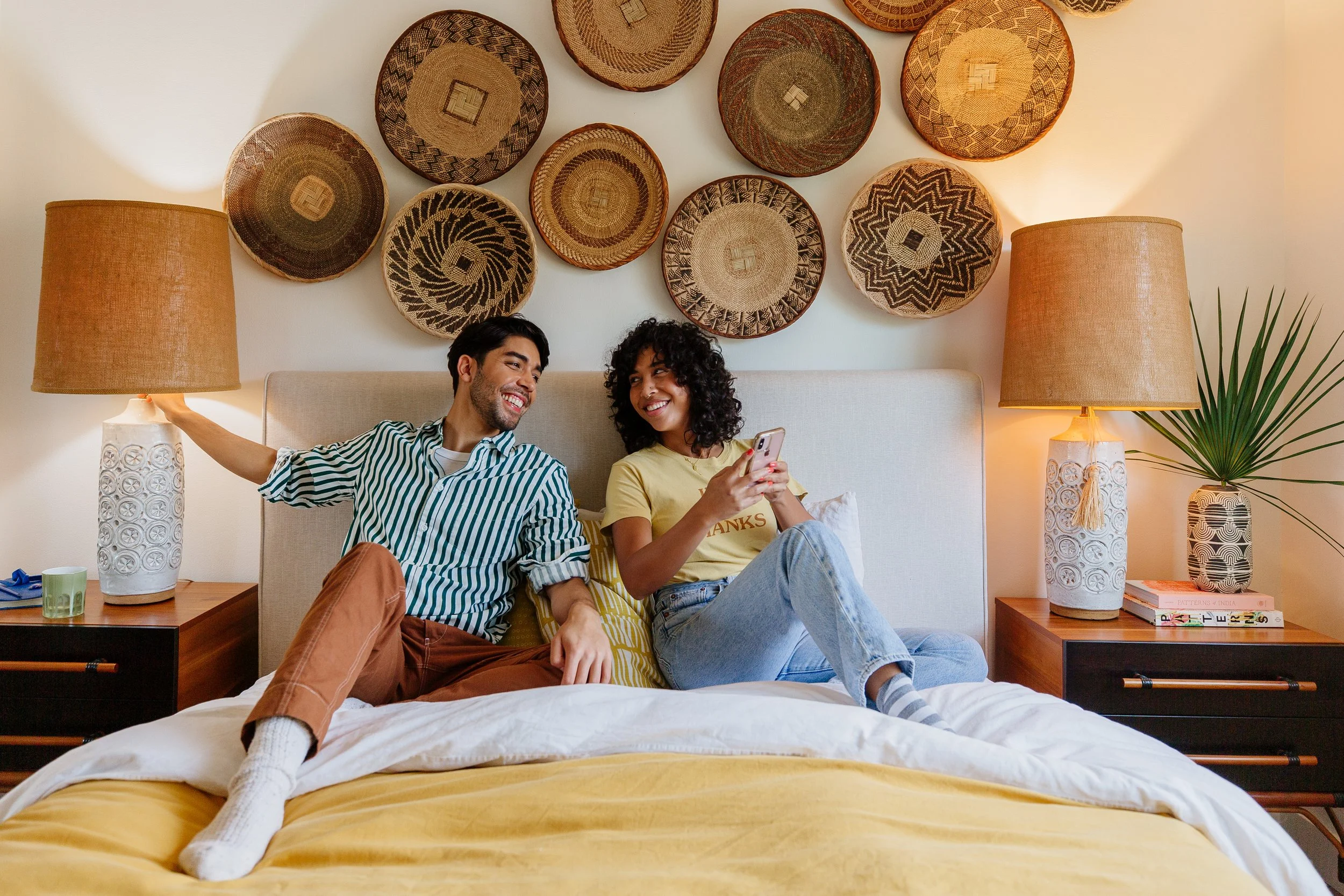 Two people sitting on a bed in a warmly lit bedroom, smiling and enjoying each other's company, with decorative woven baskets on the wall behind them and two matching bedside lamps.