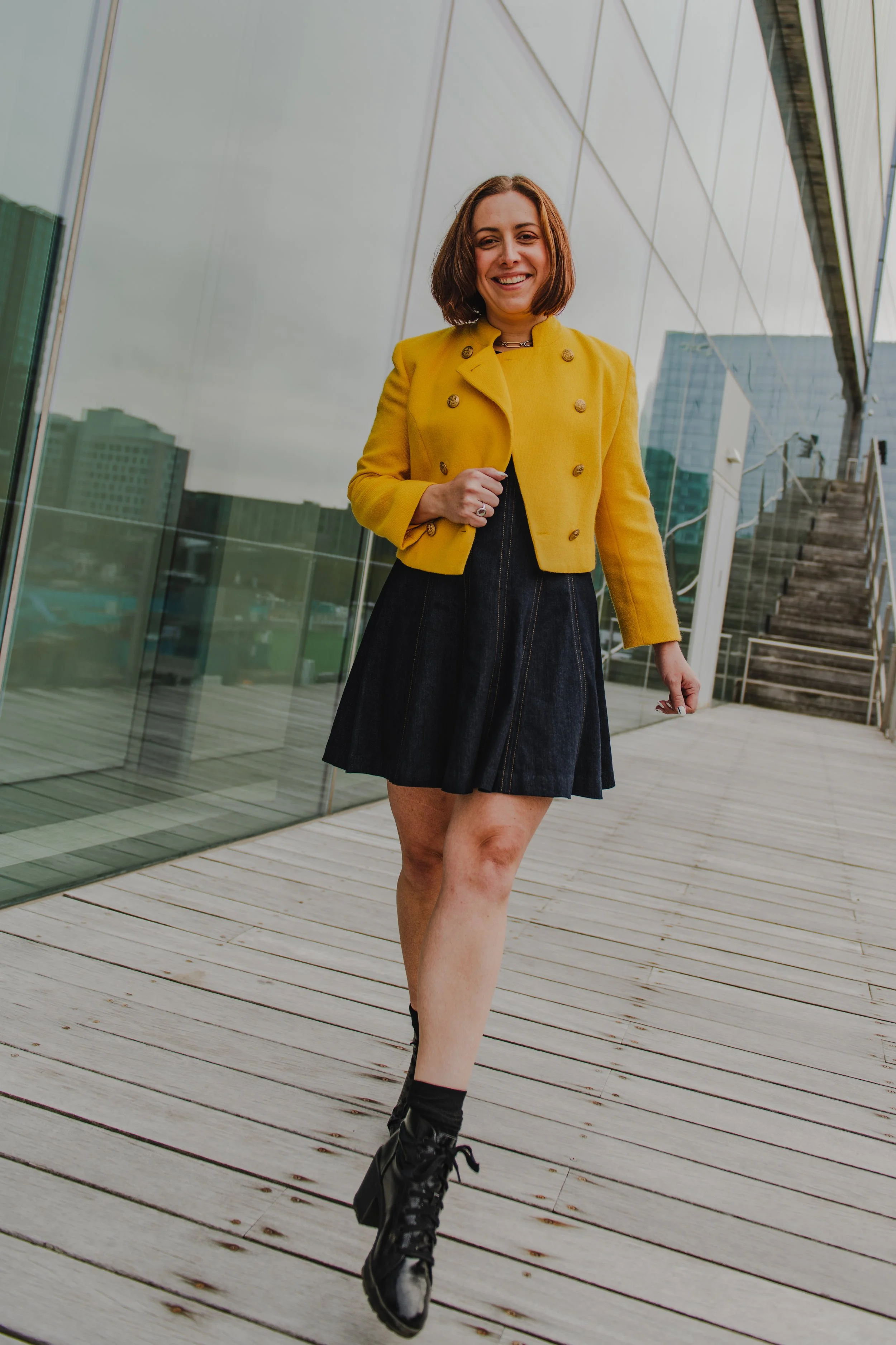 Woman with short brown hair smiling, wearing a yellow cropped jacket, black dress, and black lace-up boots, walking on a wooden deck outside a modern building with glass walls and stairs