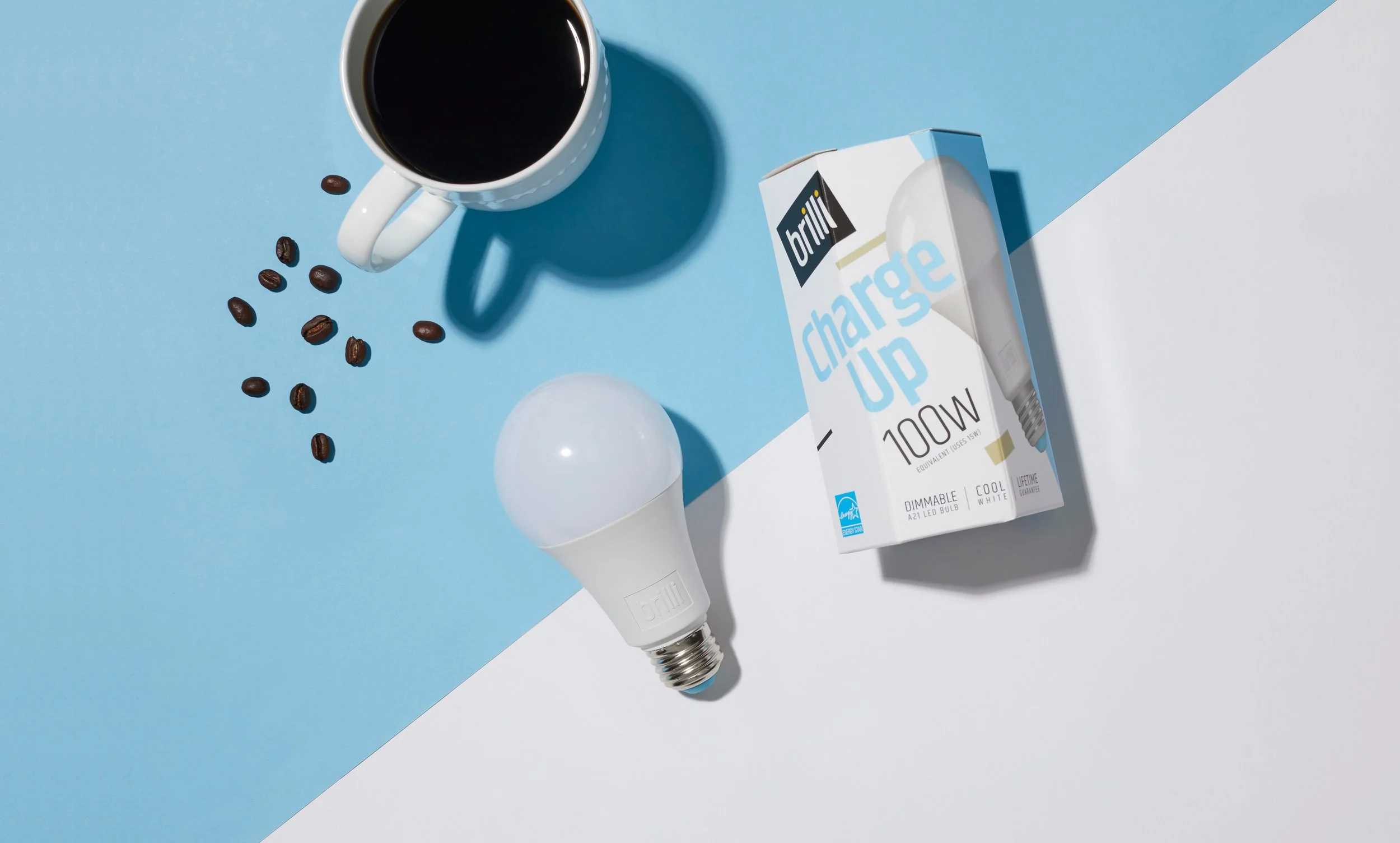 A white light bulb, a box of LED bulbs labeled charging up 100W, a cup of black coffee with coffee beans around it, and a white mug are placed on a surface divided into blue and white sections.