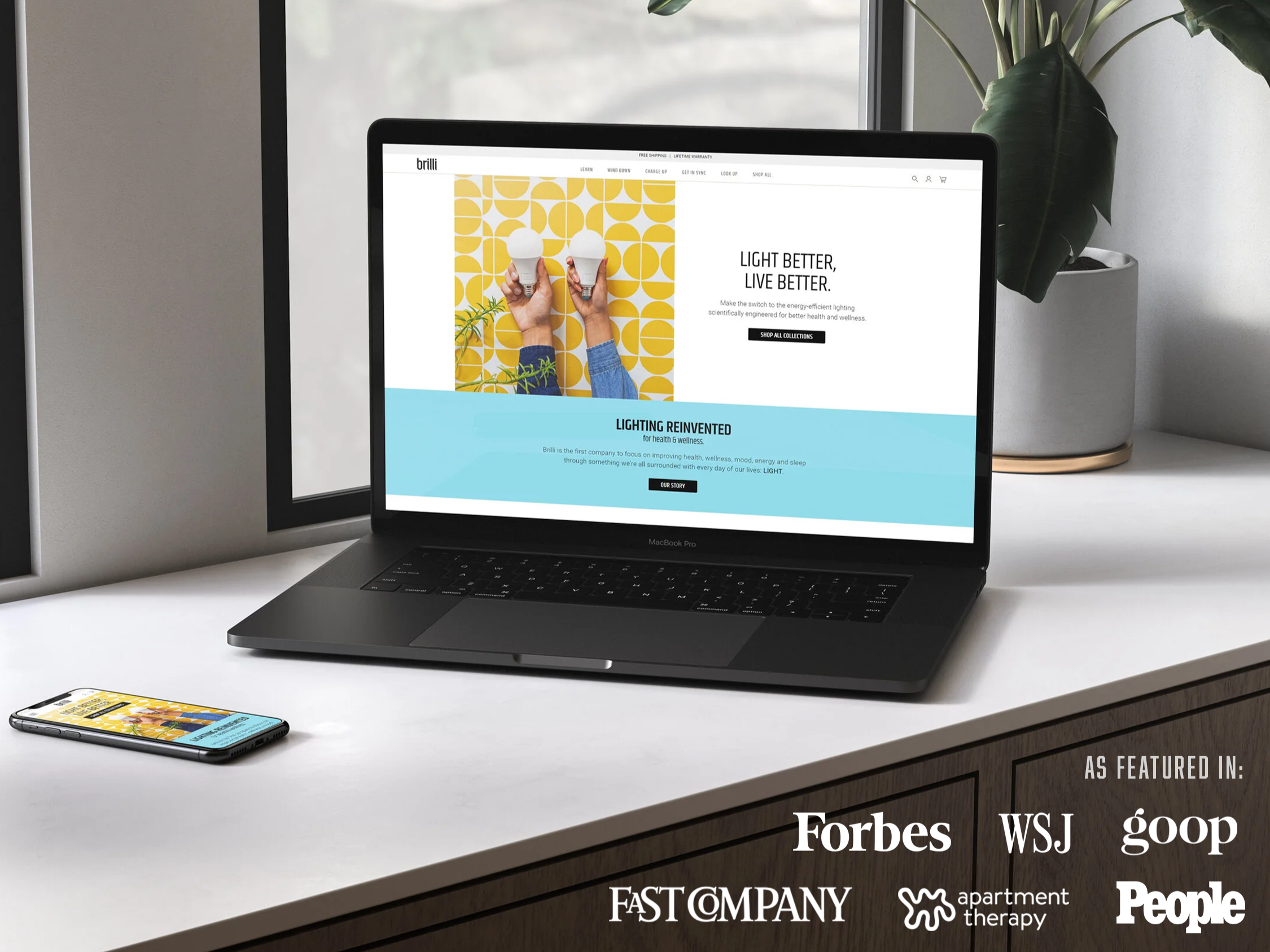A MacBook Pro on a white desk displays a webpage about energy-efficient lighting, with a smartphone nearby showing the same page. A plant in a white pot sits behind the laptop, against a window with natural light.