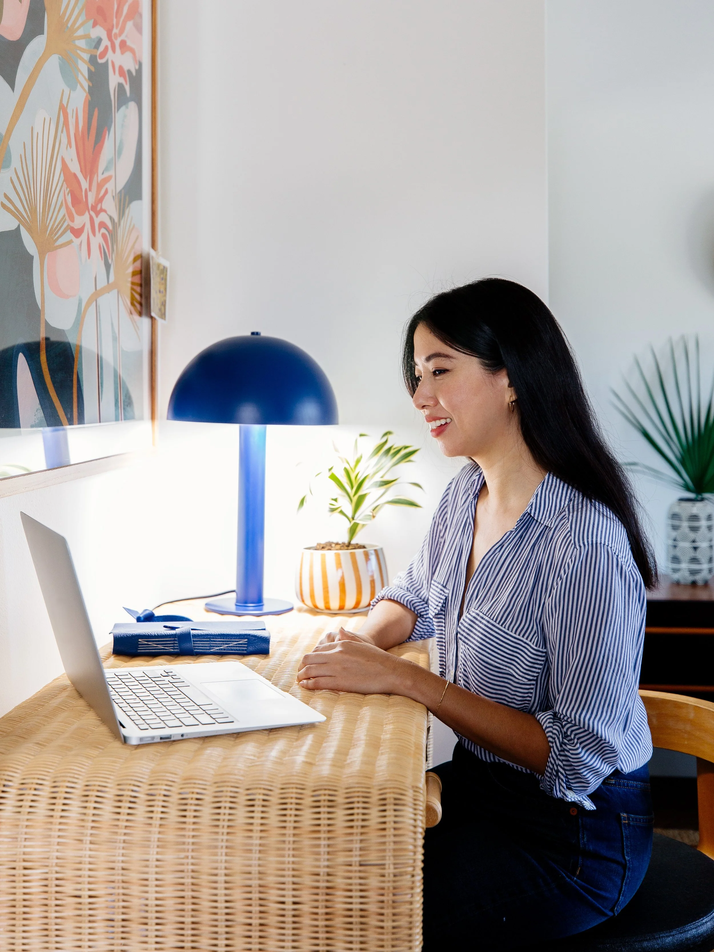 A woman sitting at a wicker table, smiling at her laptop, with a blue desk lamp and potted plants in a bright, decorated room.