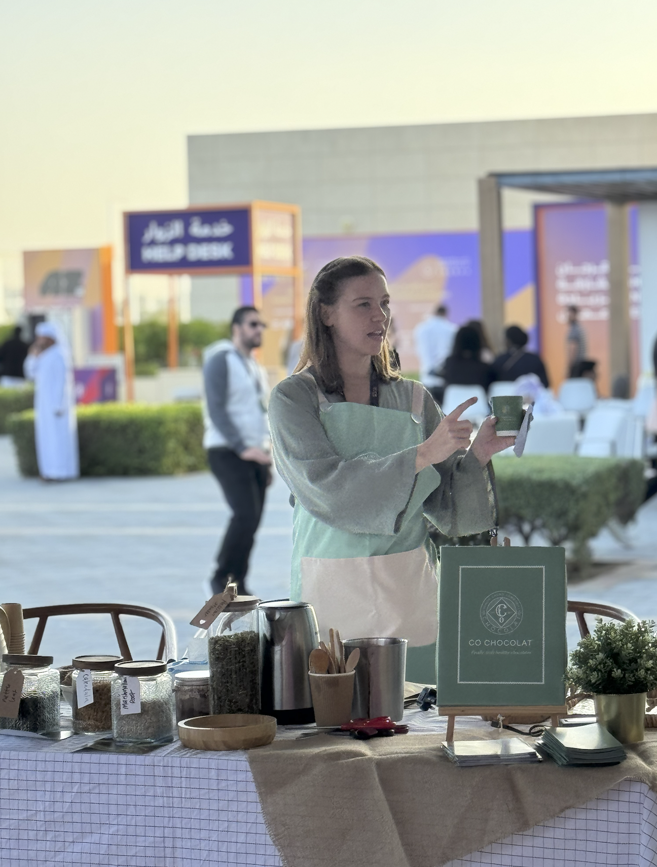 Woman at an outdoor market stall, pointing at her phone. The stall displays jars of ingredients, a sign for Co Chocolat, and various kitchen items, with people walking in the background.