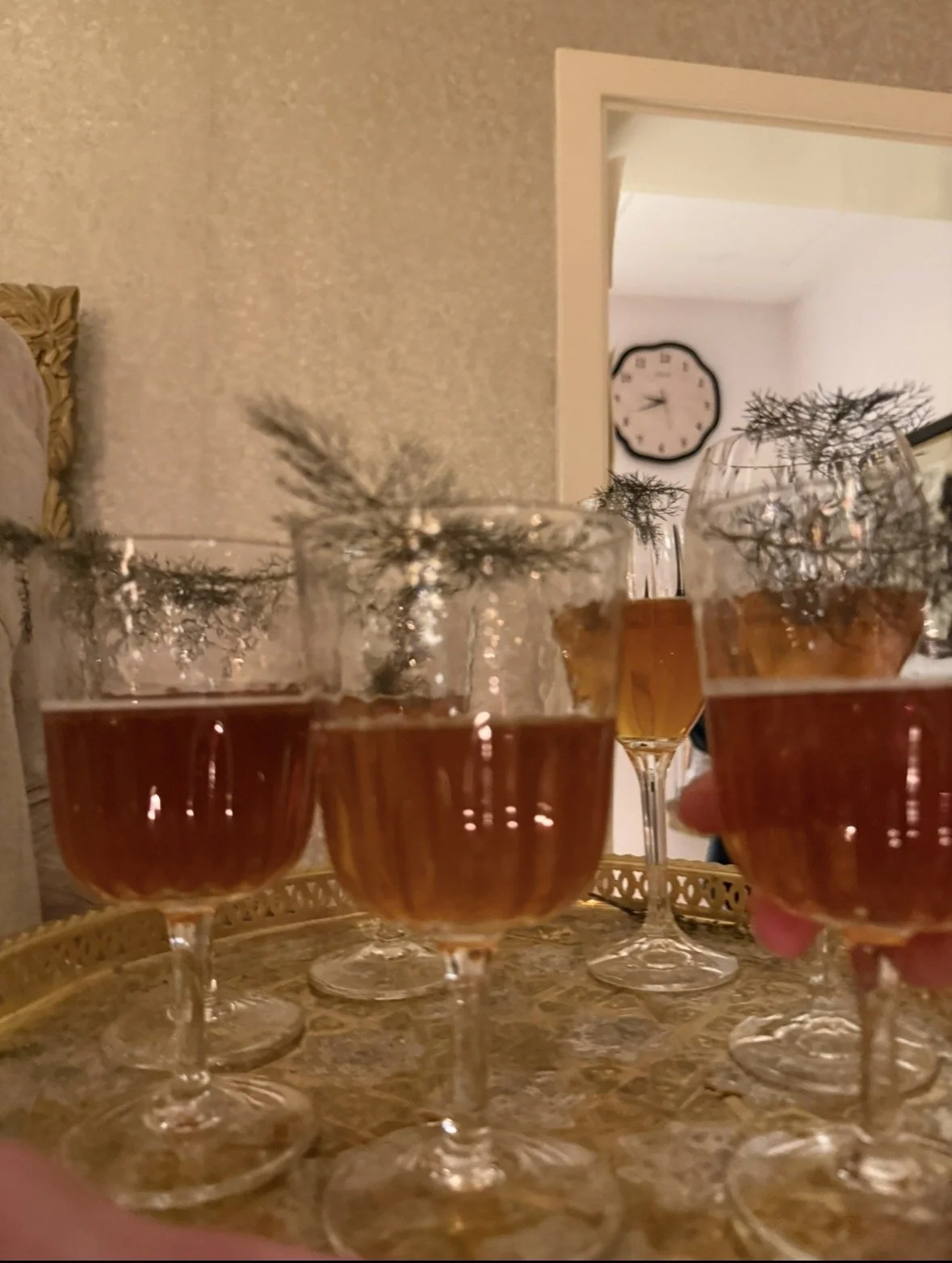 Multiple wine glasses filled with a dark red beverage on a table, with Christmas tree branches as garnish, in a cozy indoor setting with a textured wall and clock in the background.