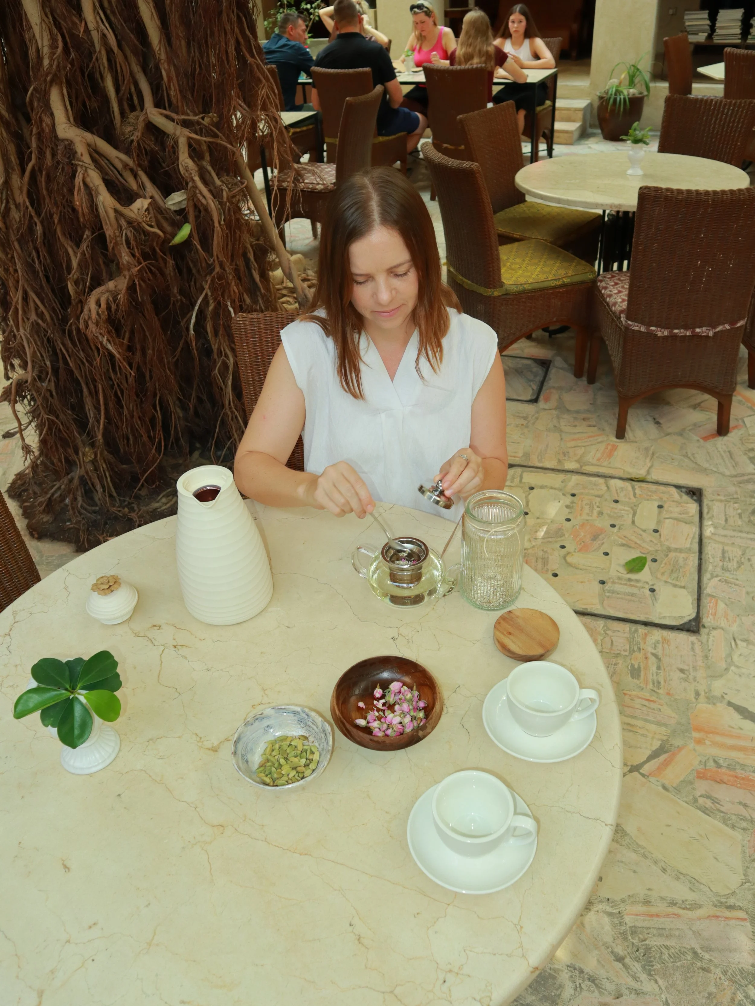 A woman sits at a round marble table with glass jars, bowls of almonds and flower petals, tea teapot, cups, and small plants, in a restaurant with people dining in the background.