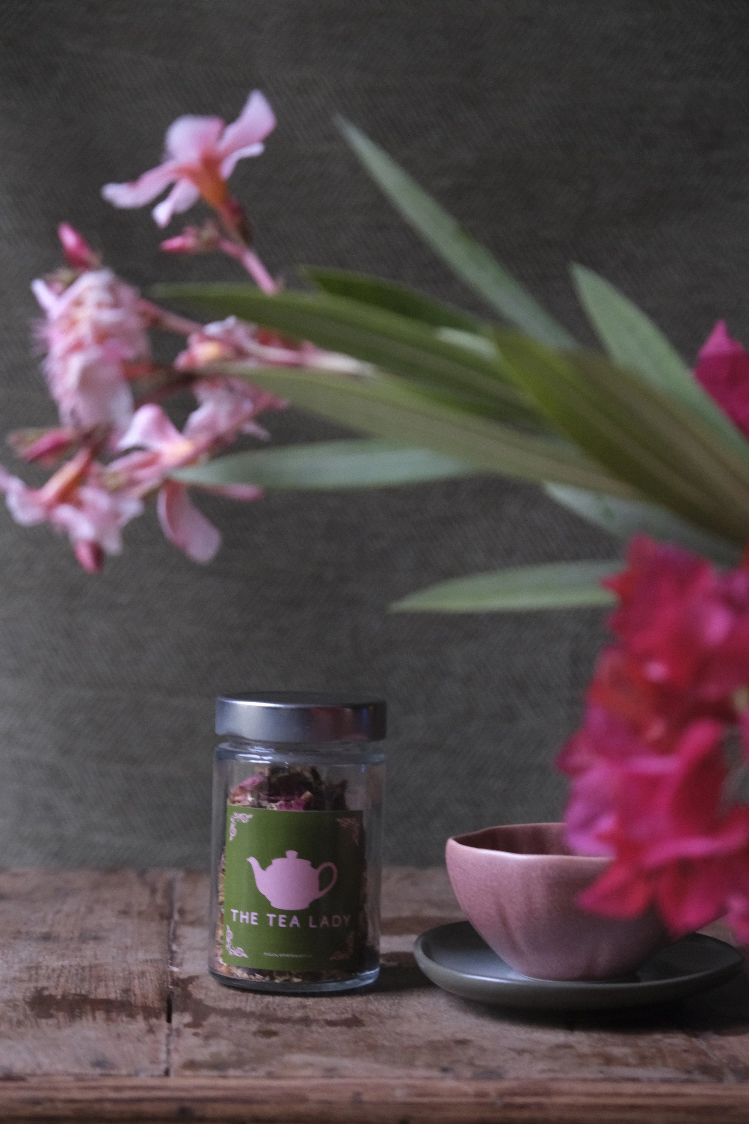 A bouquet of pink flowers with green leaves, a pink bowl with another flower, and a jar labeled "The Tea Lady" on a wooden surface with a black background.