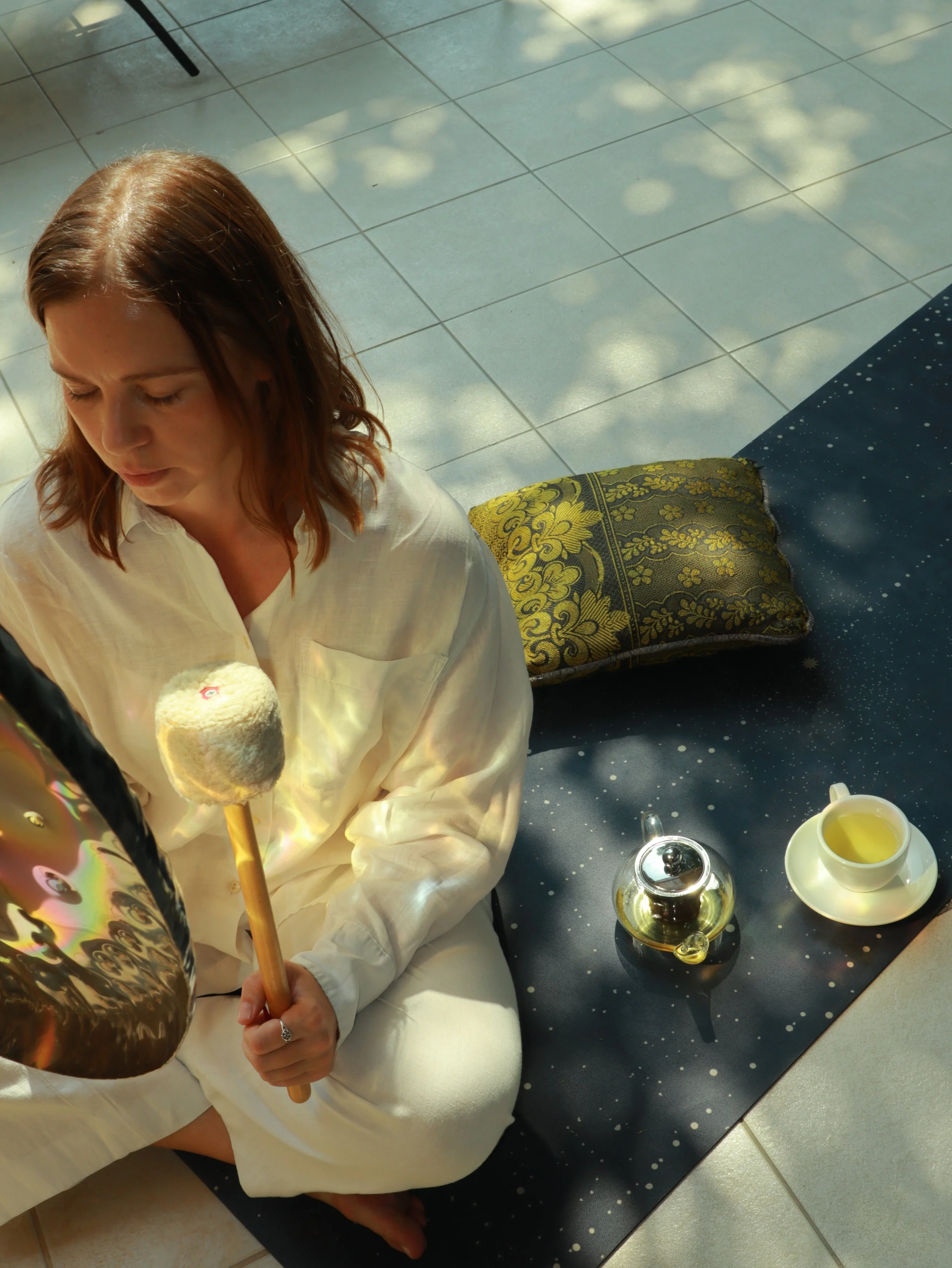 A woman with reddish-brown hair wearing a white robe sitting on the floor, holding a large, shiny balloon. There is a tea set with a teapot and a cup of yellow-green liquid on a black speckled mat beside her. Sunlight filters through the window, casting light spots on the tiled floor and on the woman and objects.