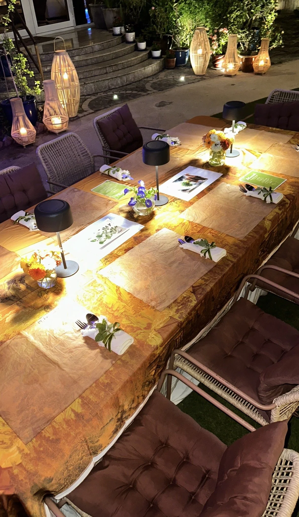 An outdoor dinner table set for a gathering with a floral centerpiece, candles, and small lamps. The table is covered with an orange tablecloth, and each place setting includes a folded napkin with a sprig of greenery. In the background, there are decorative lamps, potted plants, and a stone staircase leading to a house with glass sliding doors.