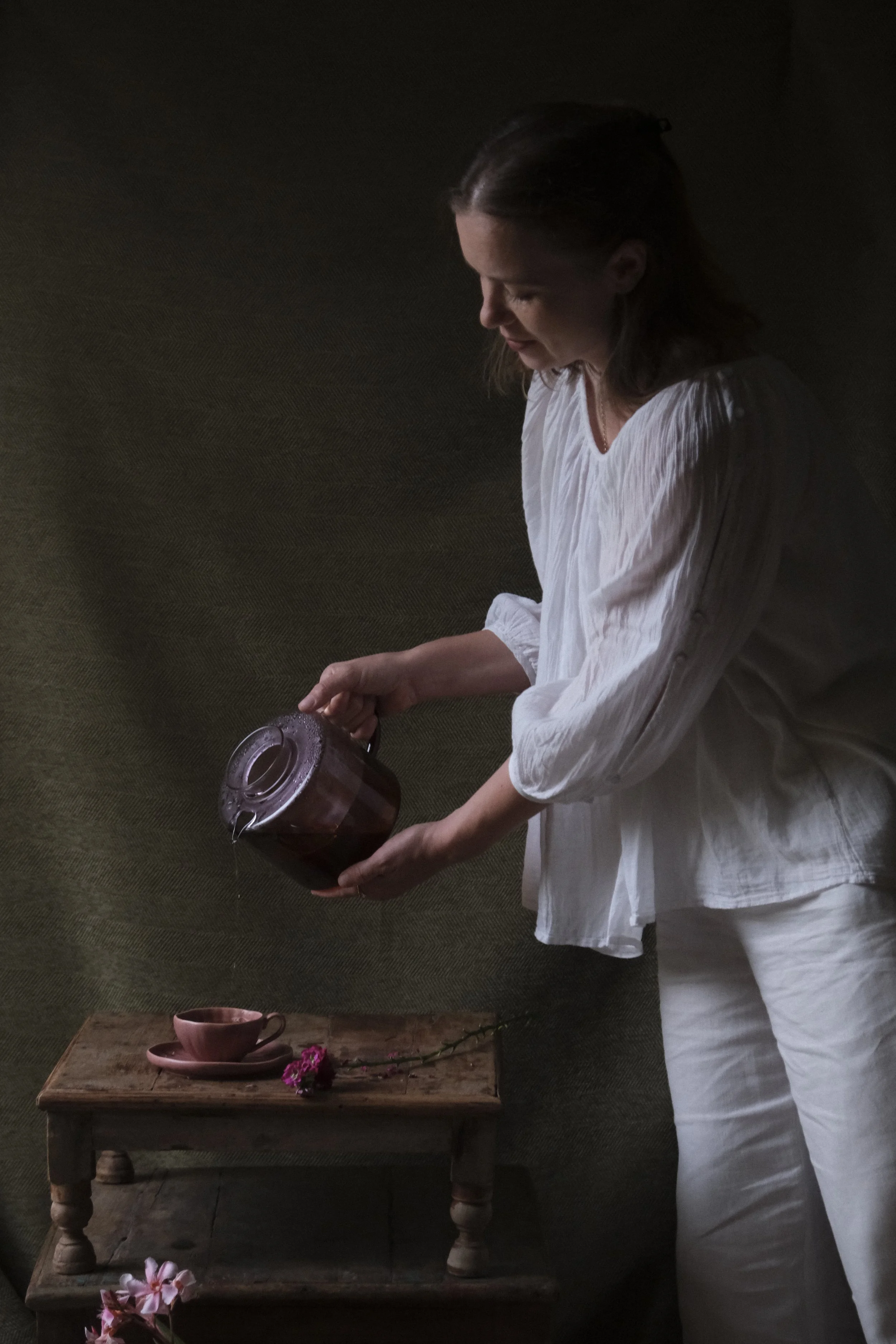 A woman in a white blouse and pajama pants pours tea from a teapot into a pink cup on a rustic wooden table. The table has pink flowers and a cup with saucer on it. The background is dark with a textured wall.