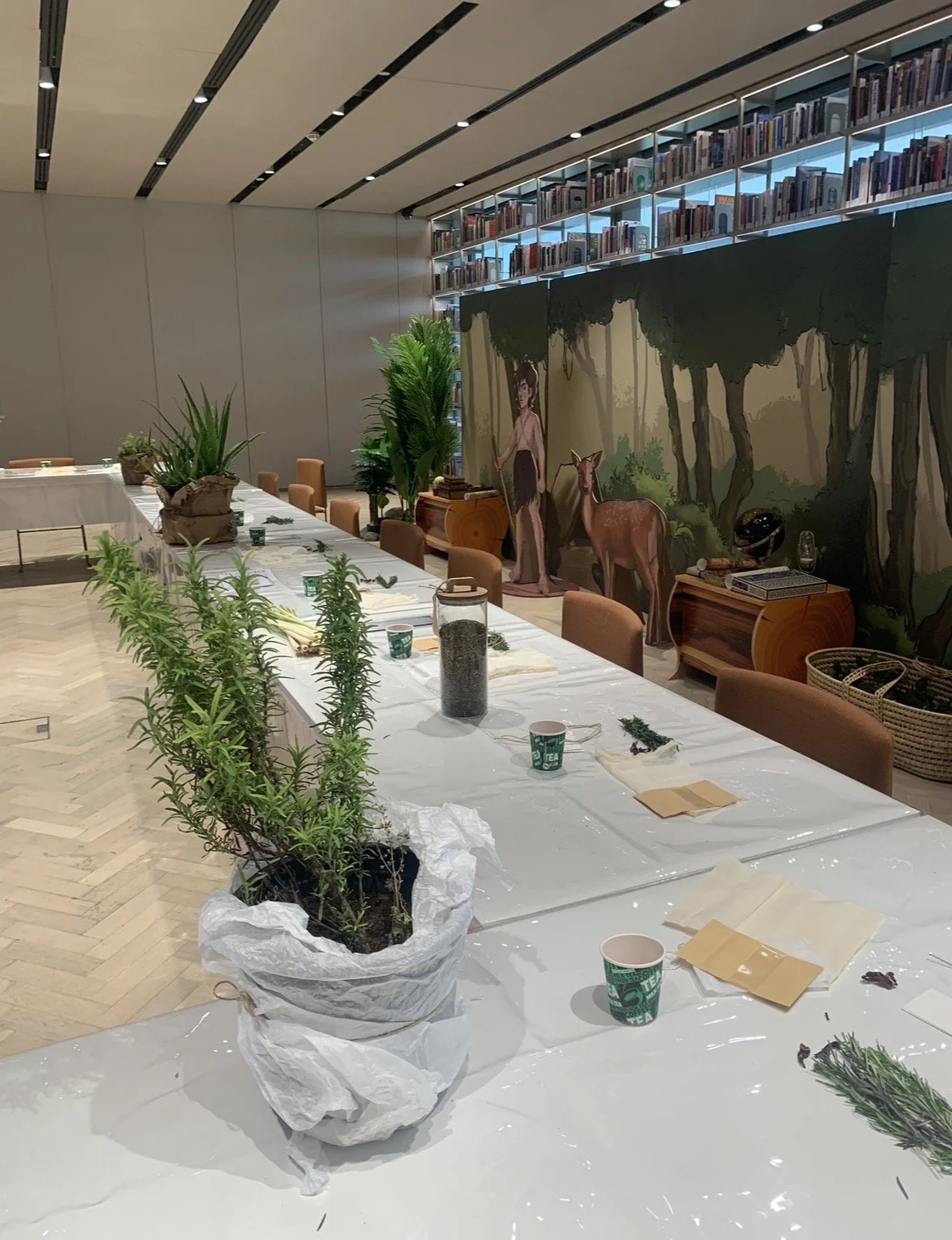 A long table set up for a plant workshop, with various plants, cups, and papers on the table. In the background, there is a mural depicting a forest scene with a woman and a deer, and shelves filled with books.