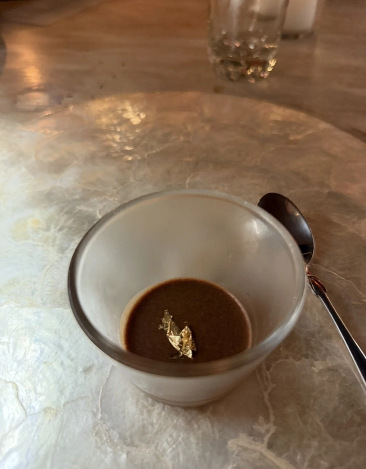 A partially used cup of black coffee with a small piece of gold foil floating on top, placed on a grayish marble table. A spoon lies next to the cup and a glass of water is in the background.