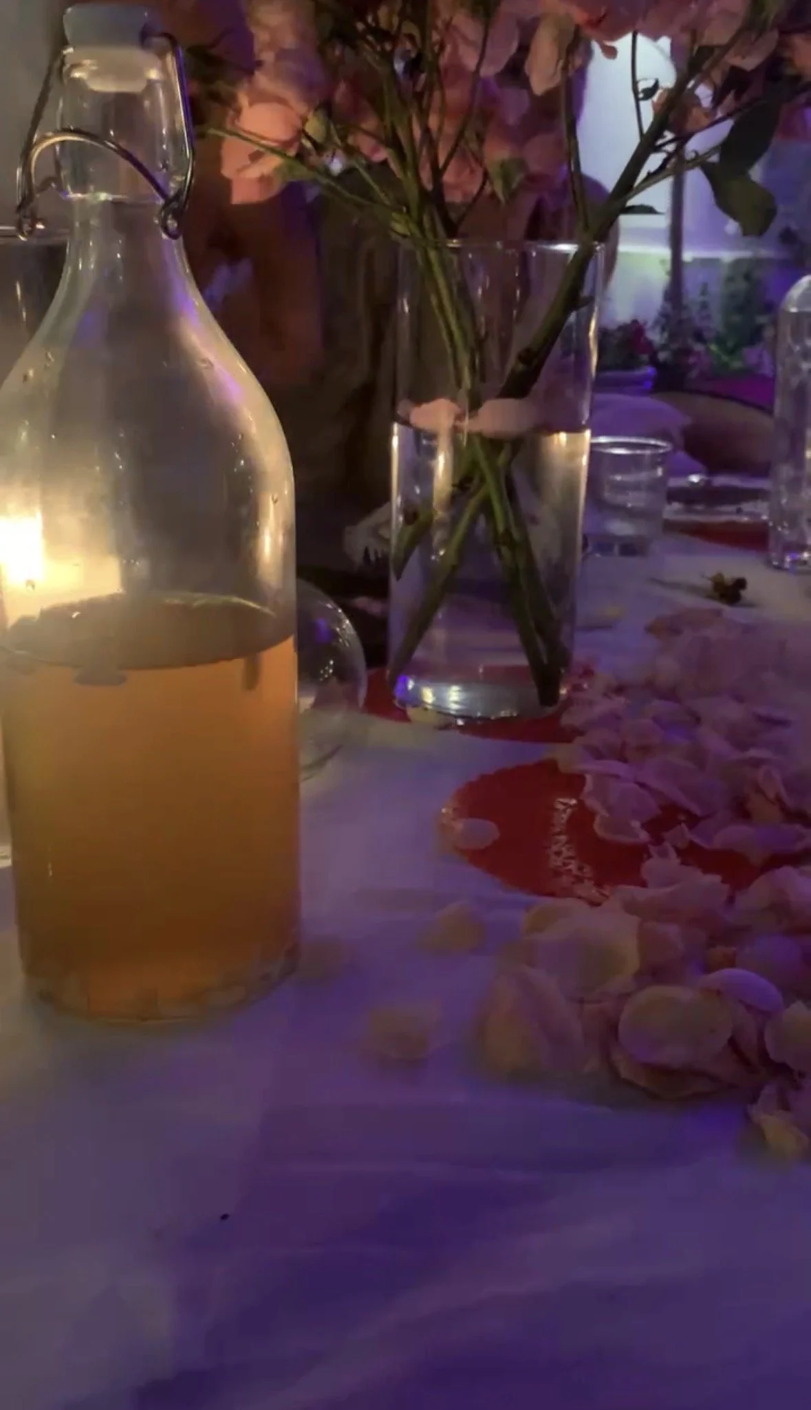 A table with a clear glass pitcher filled with a light-colored beverage, possibly juice, and a small bouquet of pink roses in a glass vase. The table appears to be decorated with white petals and is set in an elegantly lit environment with purple lighting.