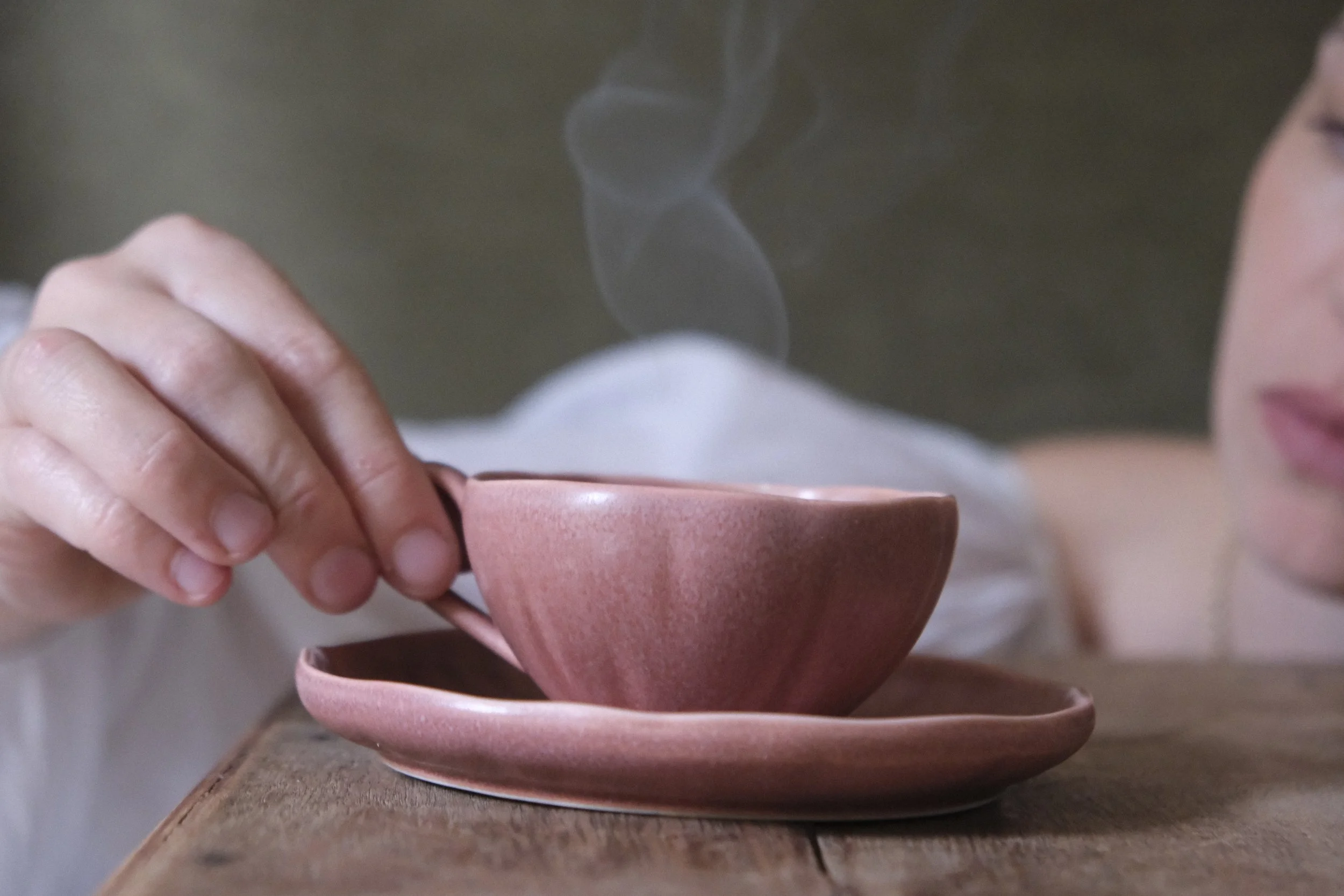 Person holding a ceramic mug over a matching saucer with a hand.