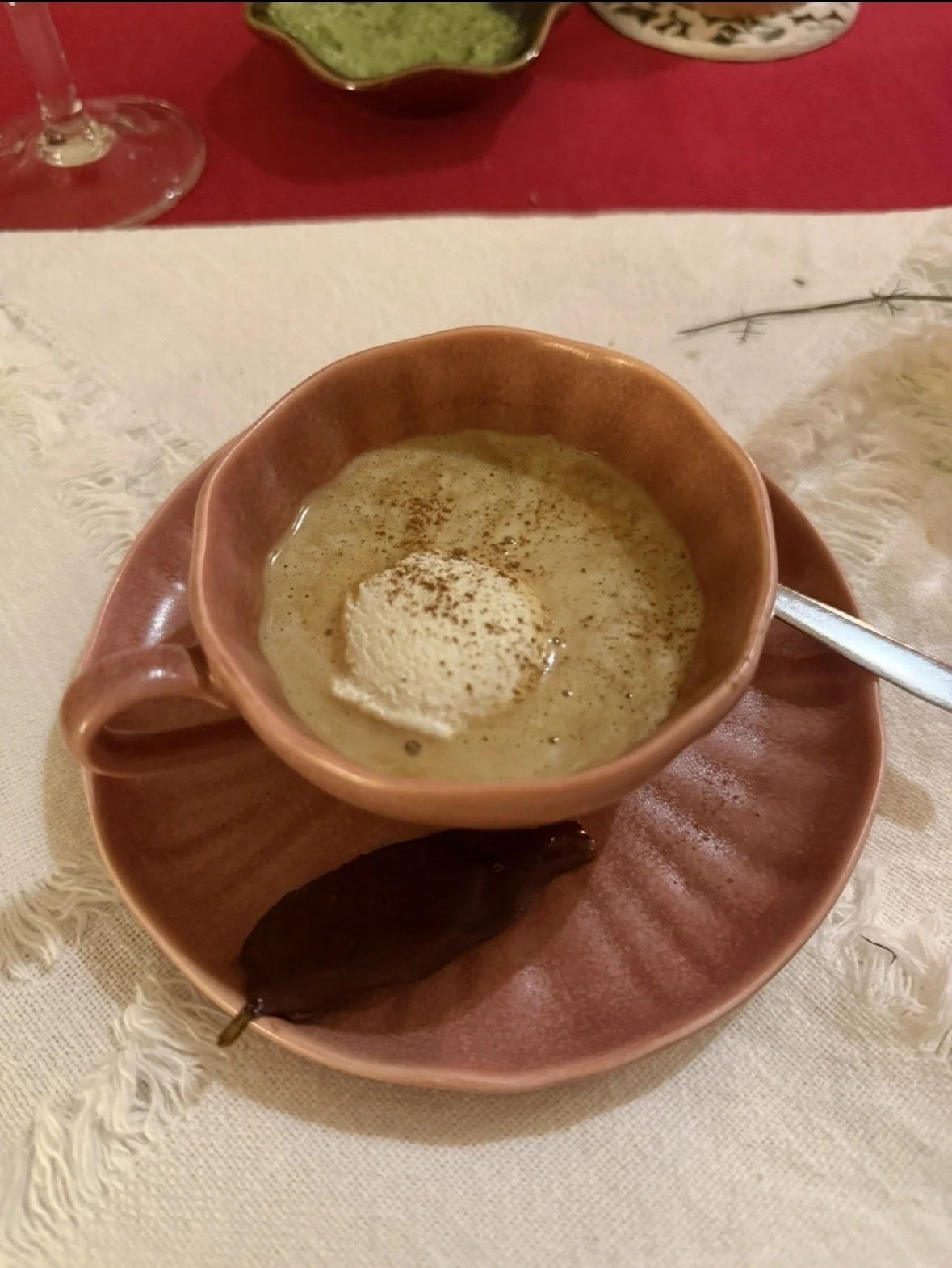 A cup of creamy soup with a dollop of sour cream or cream on top, served in a pink ceramic cup on a matching pink saucer with a spoon, placed on a white textured cloth. There is also a dark brown leaf-shaped object on the saucer.