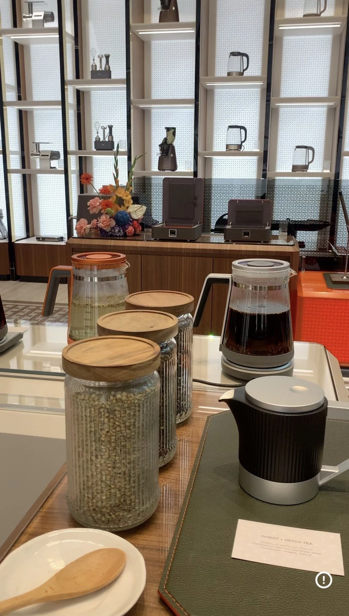 Various jars and a coffee maker on a table, with a bouquet of flowers and a menu in the background.