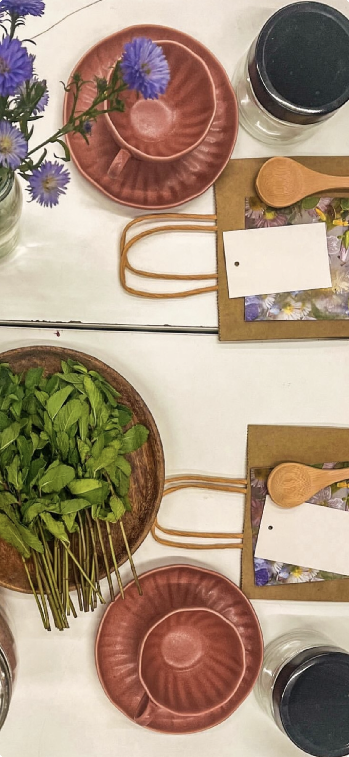 Table setting with purple flowers in a vase, a pink cup and saucer, a wooden spoon, a glass container with a black lid, and a plate of fresh green herbs.