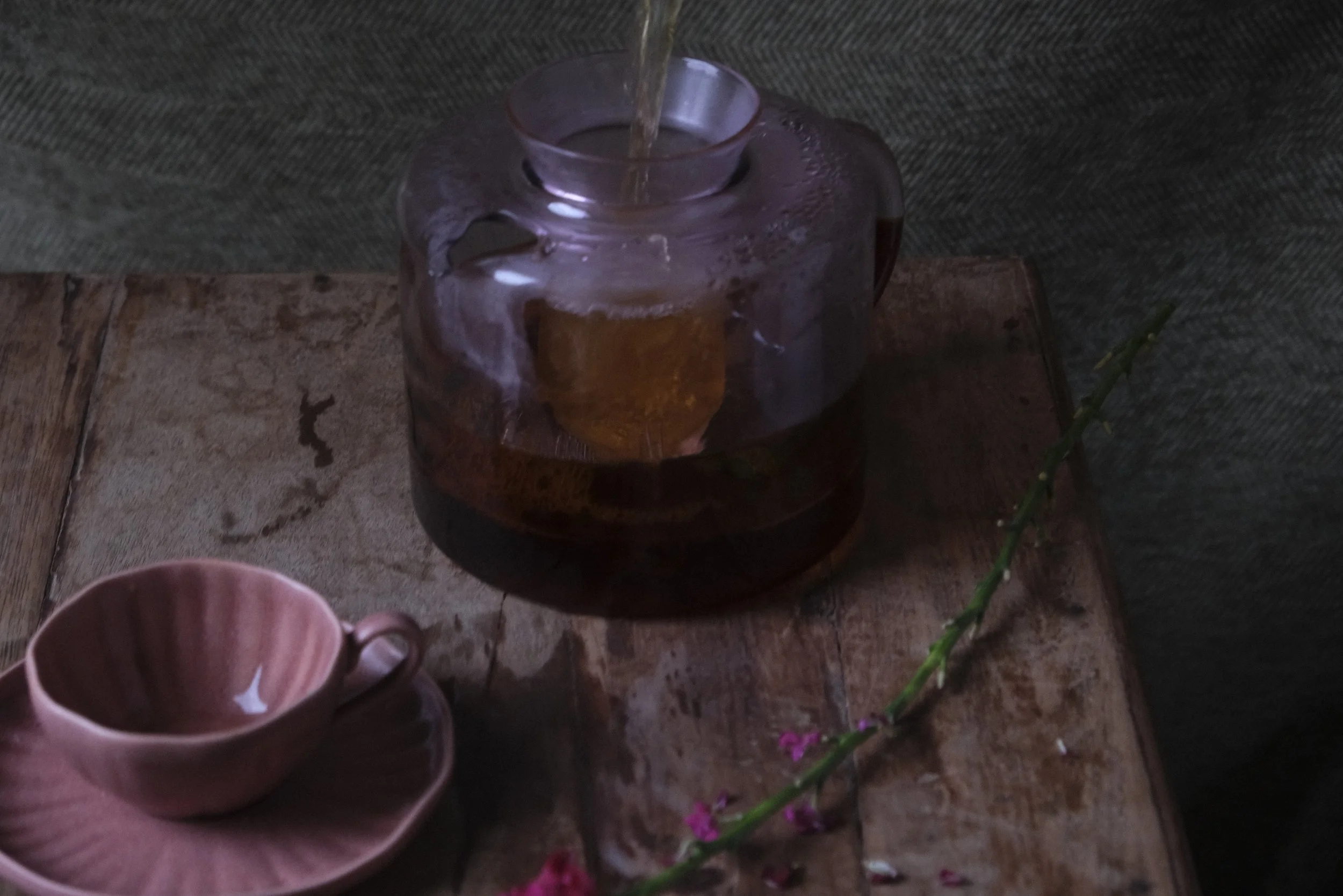 A clear glass teapot with a metal lid and a handle, placed on a wooden surface with a textured dark background. A person is holding a pink ceramic cup with a flower-shaped design. There is a thin green plant with small pink flowers in the foreground.