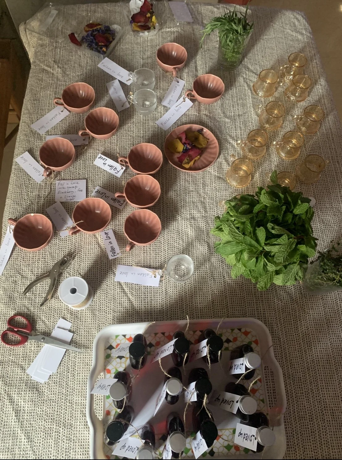 A table decorated with various items for a tea party or gathering, including pink teacups with tags, small glasses with tags, a basket of assorted wrapped candies, a potted mint plant, and a tray of small bottled drinks with tags. There are also crafting tools like scissors, a hole punch, and loose tags on the table.