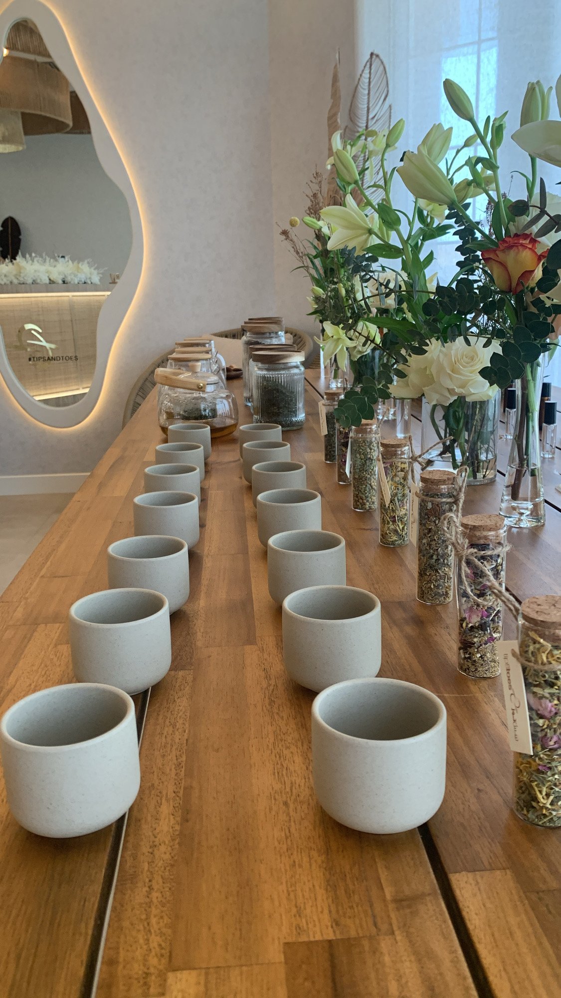 A wooden table decorated with white ceramic cups and small glass jars filled with dried herbs or flowers, alongside green leafy and white flower arrangements.