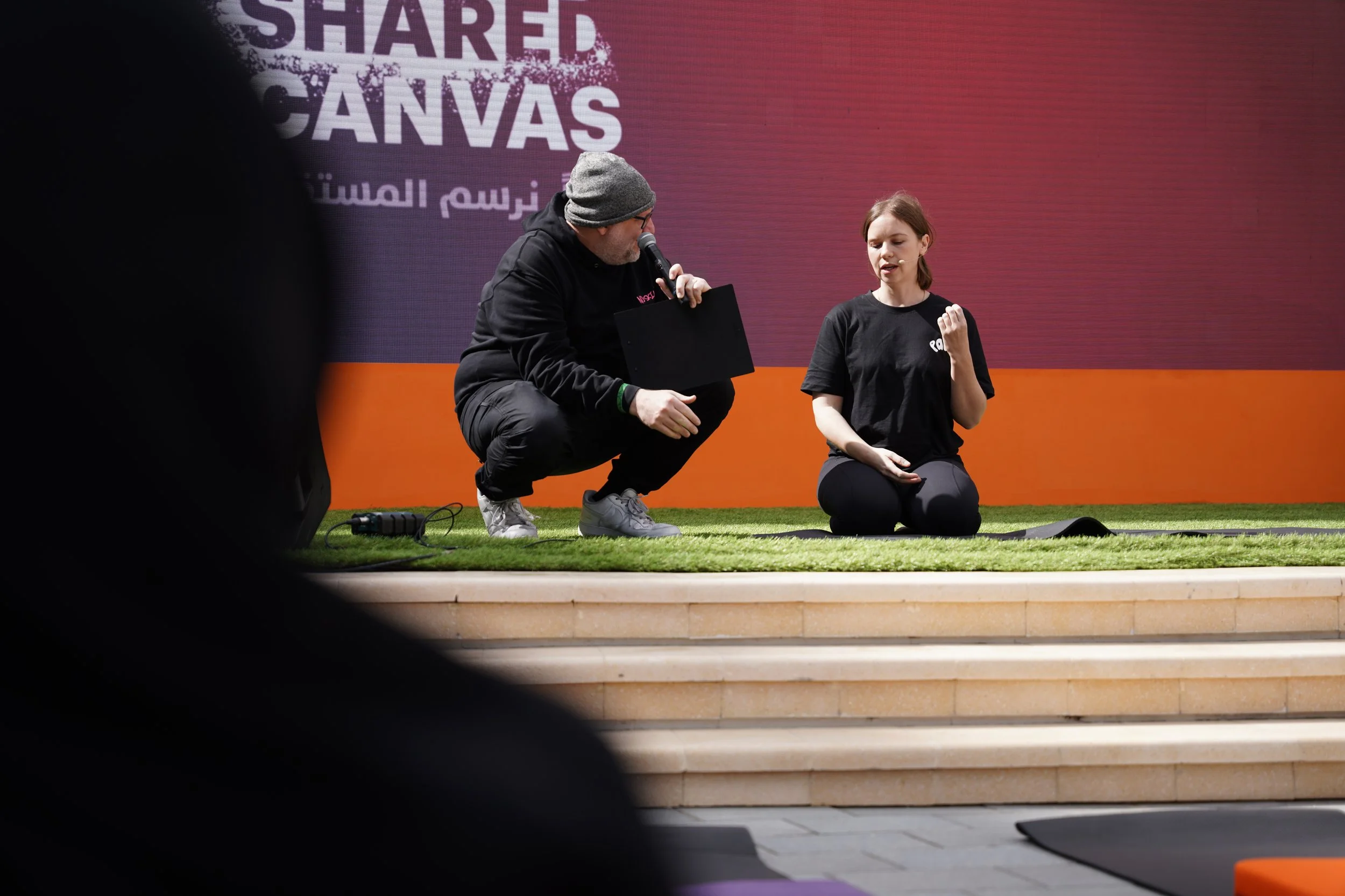 A man with glasses and a gray beanie kneeling and speaking into a microphone while holding a black tablet, and a woman with brown hair and a black t-shirt sitting on the stage with her fist raised, in front of a purple and orange background with partially visible text.