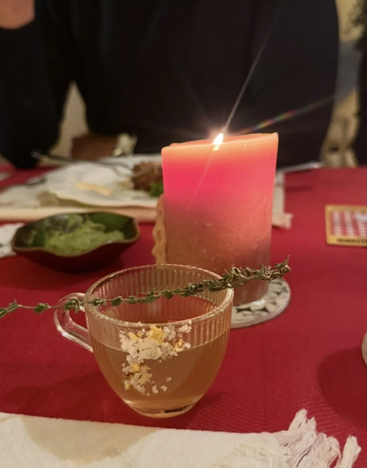 A candle and a glass of drink on a dinner table with food dishes in the background, some people seated at the table, a red tablecloth, and a sprig of herbs.